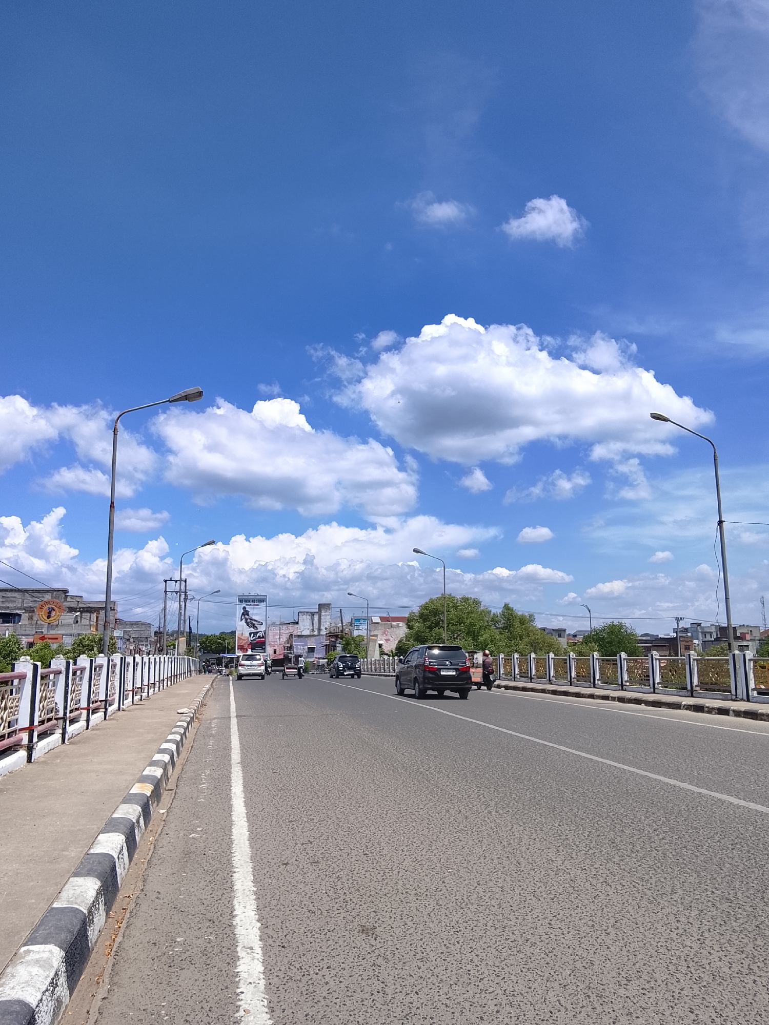 The image depicts a street scene in Lhokseumawe, Indonesia, on a clear day with a bright blue sky and scattered white clouds. A two-lane asphalt road runs through the center of the frame, with a pedestrian walkway and decorative railing along the left side. Several vehicles, including cars and a scooter, are visible on the road. Buildings, trees, and streetlights line the sides of the road. The overall impression is of a typical urban environment on a sunny day.