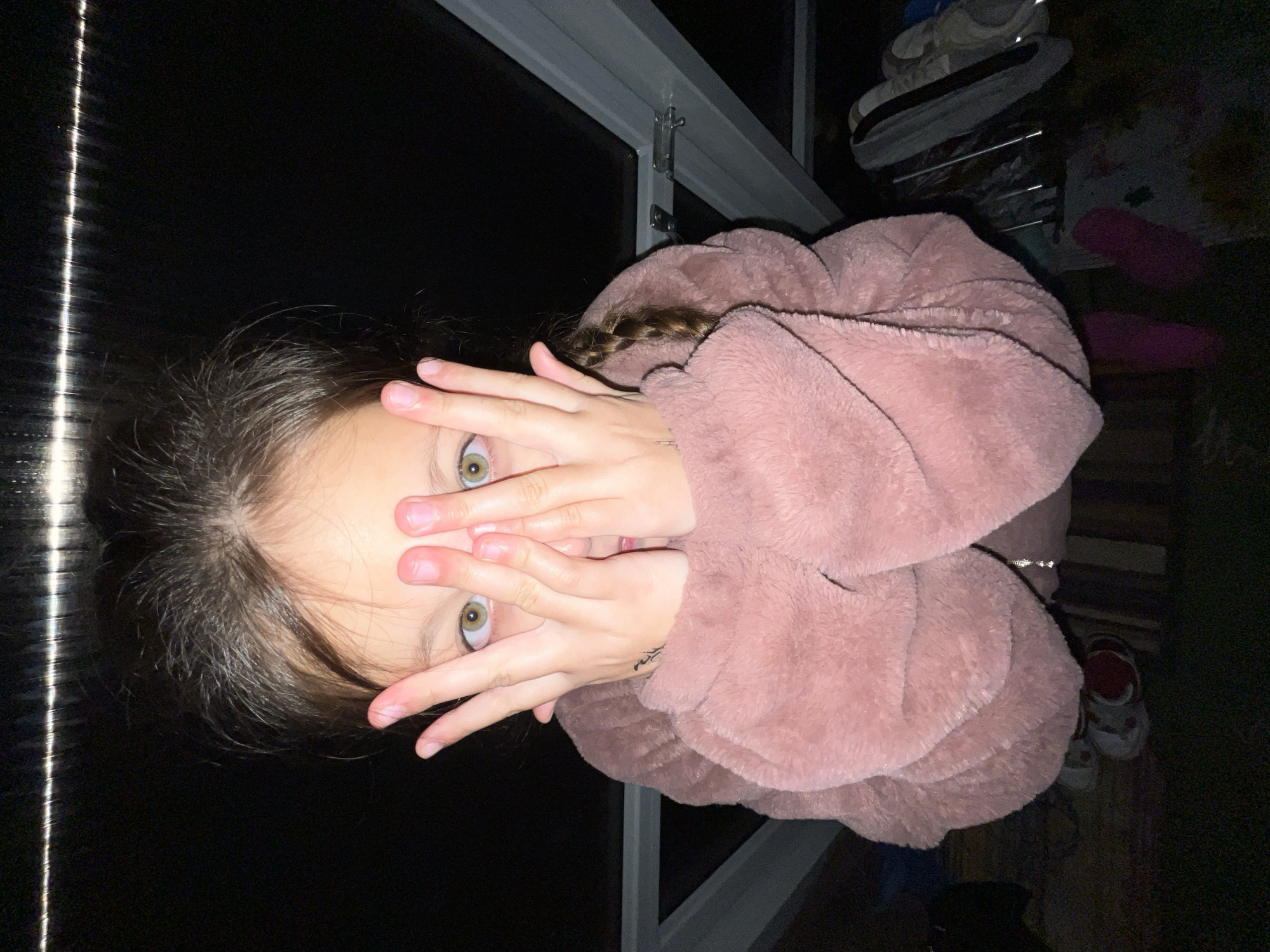 A girl with light brown hair and green eyes covers her face with her hands, peering through her fingers. She wears a light pink, fluffy jacket. She has a braid visible at the side of her head. The background is dark and includes a white metal window frame and blurred outlines of shoes and other objects. The location is not identifiable from the image.