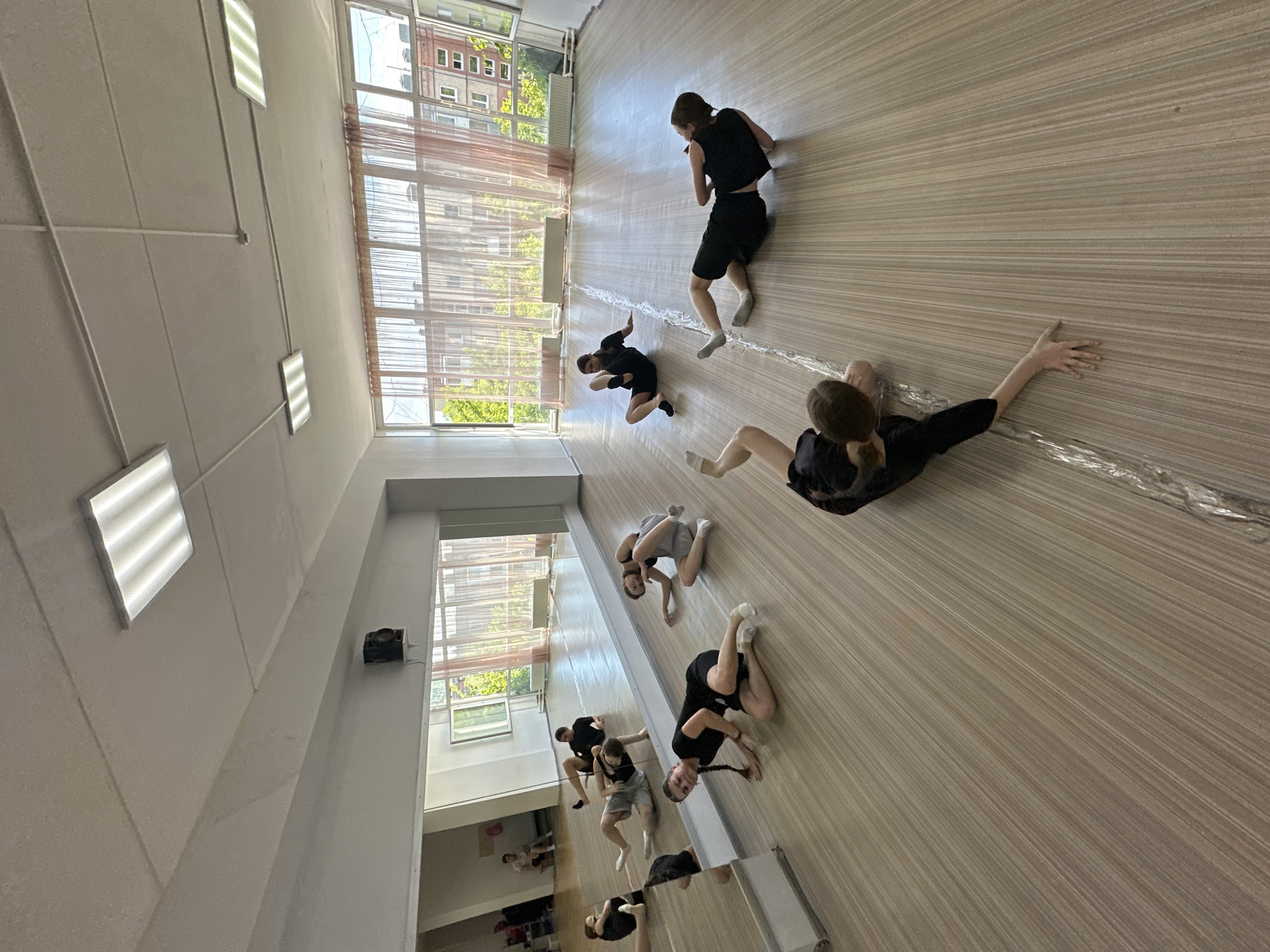 The image depicts a group of young people, likely students, engaged in a dance or movement class in a studio. The studio has a large mirrored wall on the left and floor-to-ceiling windows with sheer pink curtains on the right, revealing an urban exterior with multi-story buildings and greenery. The floor is made of light-colored wood planks, and fluorescent lights are mounted on the ceiling.
Several individuals are positioned on the floor in various dynamic poses, suggesting they are in the midst of a choreographed sequence or a stretching exercise. Most are wearing dark athletic clothing, primarily black tops and shorts or leggings, and white socks.
In the foreground, a person is lying on their side, reaching out an arm. Further back and to the right, another person is on their hands and knees. More individuals are scattered across the room, some in seated or kneeling positions, and others in more complex floor-work poses. The reflection in the mirror shows additional people, some seated and observing.
The overall atmosphere is one of focused activity within a dedicated practice space. The image was captured in the city of Votkinsk, Russia.