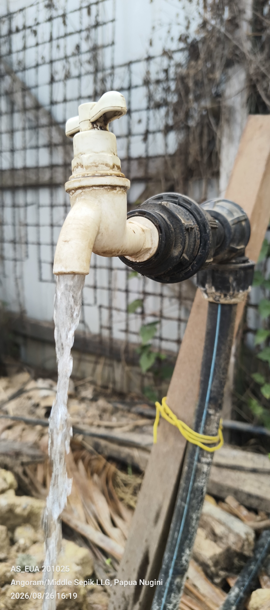 A close-up, vertical shot captures a white plastic water faucet actively dispensing a stream of water. The faucet is connected to a black pipe fitting. In the background, a white, wire-mesh-like fence is visible, with dry, brown foliage and some green plants interspersed. To the right, a rough-hewn wooden beam leans diagonally, secured by a bright yellow elastic band. The ground is covered with dry leaves and debris. The lower-left corner of the image contains text overlays with the following information: "AS_EUA 201025", "Angoram/Middle Sepik LLG, Papua Nugini", and "2026/08/26 16:19". The image appears to have been taken outdoors, likely in a rustic or undeveloped area.