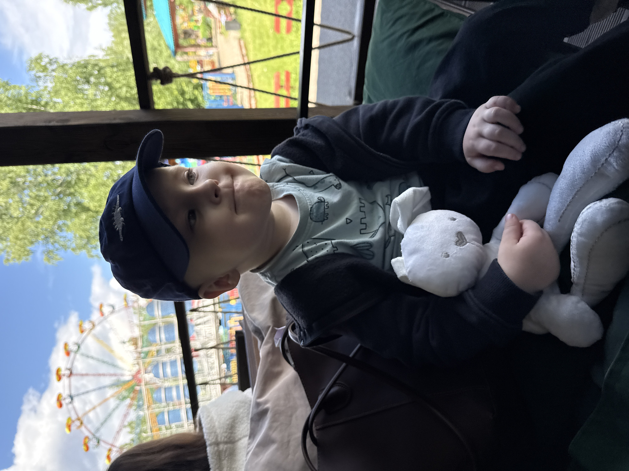 A young child wearing a navy blue baseball cap and a light blue patterned shirt is holding a white stuffed bunny rabbit. The child is sitting, seemingly on a dark green cushion. In the background, there is a Ferris wheel with colorful gondolas, indicating an amusement park setting. The child and the Ferris wheel are viewed through a partially obstructed window or opening. The location is identified as the city of Chaikovsky, Russia. The exact position within the city and the actions occurring, beyond the child sitting and holding the toy, are unclear from the image.