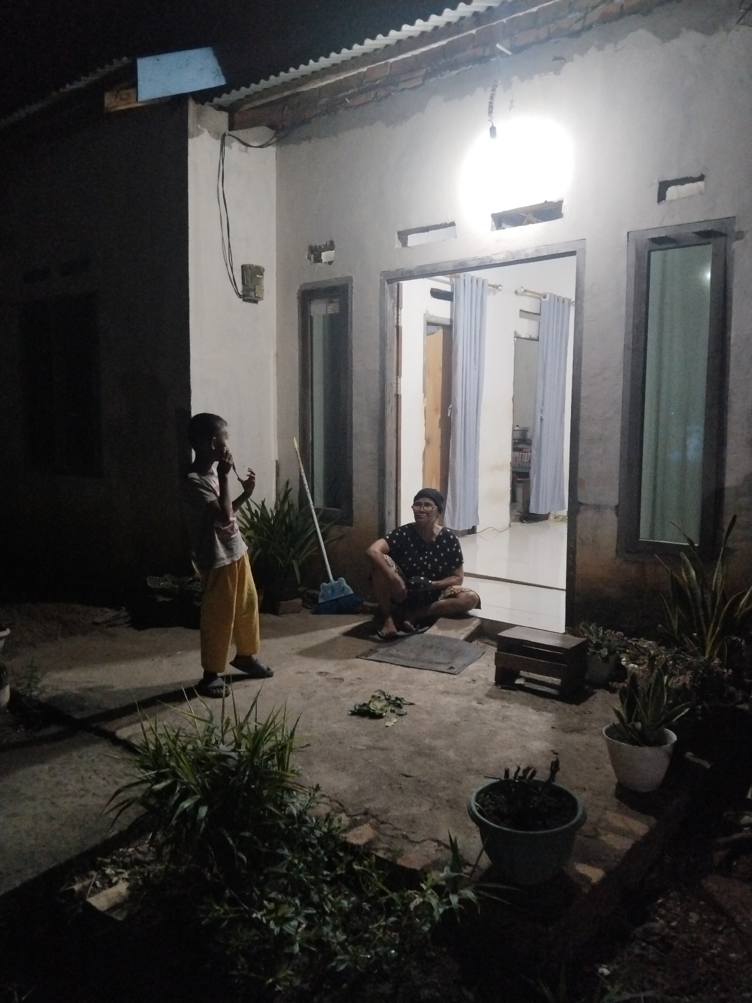 The image depicts an outdoor patio area at night, likely at a residence in Suka Ramai, Indonesia. A young boy stands near a wall on the left, wearing a light-colored t-shirt and yellow pants. He appears to be holding something to his mouth, possibly food or a drink. In the doorway of the house on the right, a person wearing glasses and a dark, patterned t-shirt with shorts is seated on the ground. The doorway is open, revealing a well-lit interior with curtains. The patio is paved, with various potted plants scattered around. A broom leans against the wall near the boy, and a small wooden stool is placed in front of the doorway. The overall lighting is dim, with a bright outdoor light illuminating the entrance.
