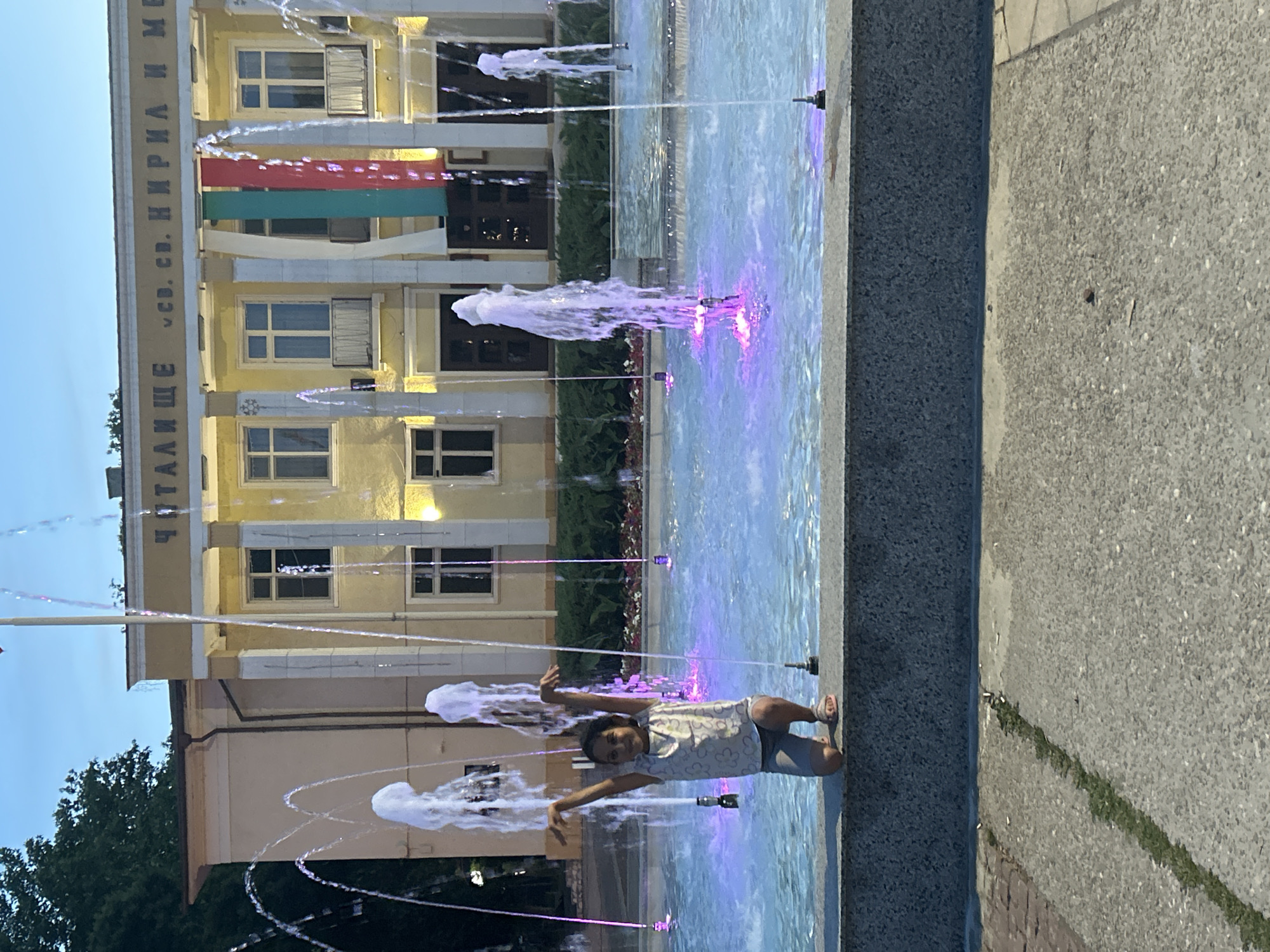 In Parvomay, Bulgaria, a young girl with dark hair is kneeling on the edge of a public fountain. She is wearing a light-colored patterned t-shirt and grey shorts, with beige sandals on her feet. Her arms are raised in the air, and she is looking towards the camera with a smile. The fountain's water is illuminated with purple and blue lights, creating a vibrant display. In the background, a yellow building with white columns stands prominently. The building has a sign with Cyrillic writing and features Bulgarian flags hanging from its facade. Trees are visible to the left of the building. The ground in front of the fountain is paved with concrete, with a line of grass growing in a crack. The scene appears to be a public square or park.