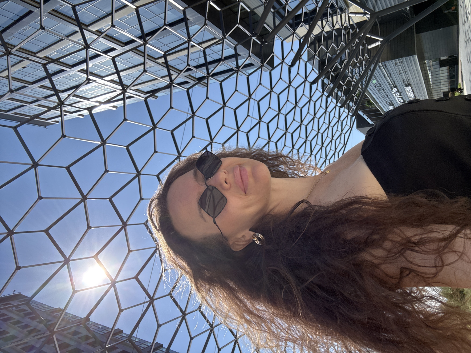 In Moscow, Russia, a woman with long, dark, curly hair stands outside under a modern, geometric metal canopy. She is wearing sunglasses with a diamond-shaped frame and a black top. Behind her, the bright blue sky is visible through the canopy's hexagonal pattern, and the sun glints brightly. To the right, a contemporary glass-fronted building with multiple windows rises into the sky. The building's facade appears to be a reflective glass and metal structure. The overall scene suggests an urban environment with a focus on modern architecture and natural light.