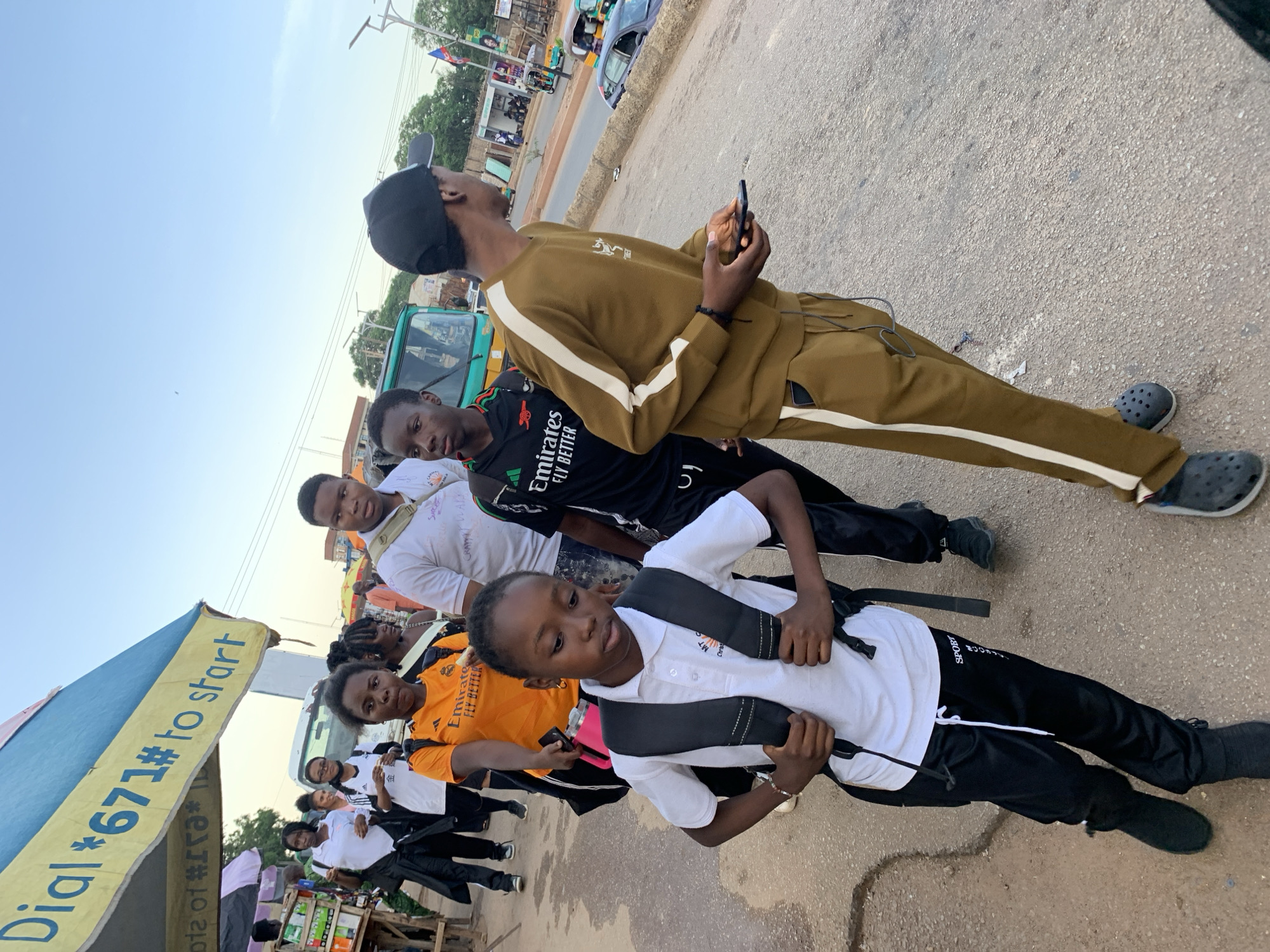 A group of young people, likely students, are walking on a paved road in what appears to be an outdoor market area. They are dressed in casual attire, with some wearing athletic clothing. A young girl in the foreground is wearing a white polo shirt with a backpack and black pants. Behind her, a person in an orange shirt and another in a black shirt are visible. Further back, a man in a brown tracksuit is walking and holding a smartphone. The environment suggests a busy urban setting, possibly Jos, Nigeria, with a blue and yellow awning overhead and various stalls and vehicles in the background. The lighting indicates it is daytime.
