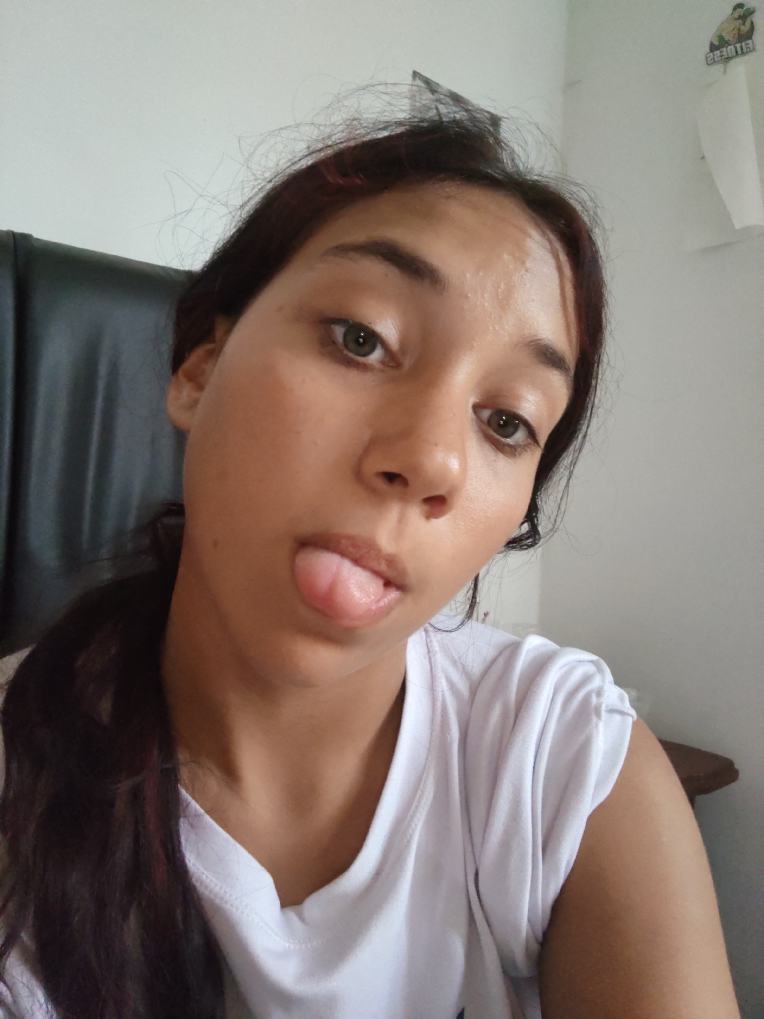 A close-up selfie shows a young woman with dark hair and brown eyes sticking her tongue out. She is wearing a white t-shirt. The background is a plain white wall. There is a sticker of a muscular figure on the wall to the right of the woman's head. A black chair is visible behind her left shoulder. The image was taken indoors, likely in a residential setting, and it is not possible to determine the specific location within Piloncito, Venezuela.