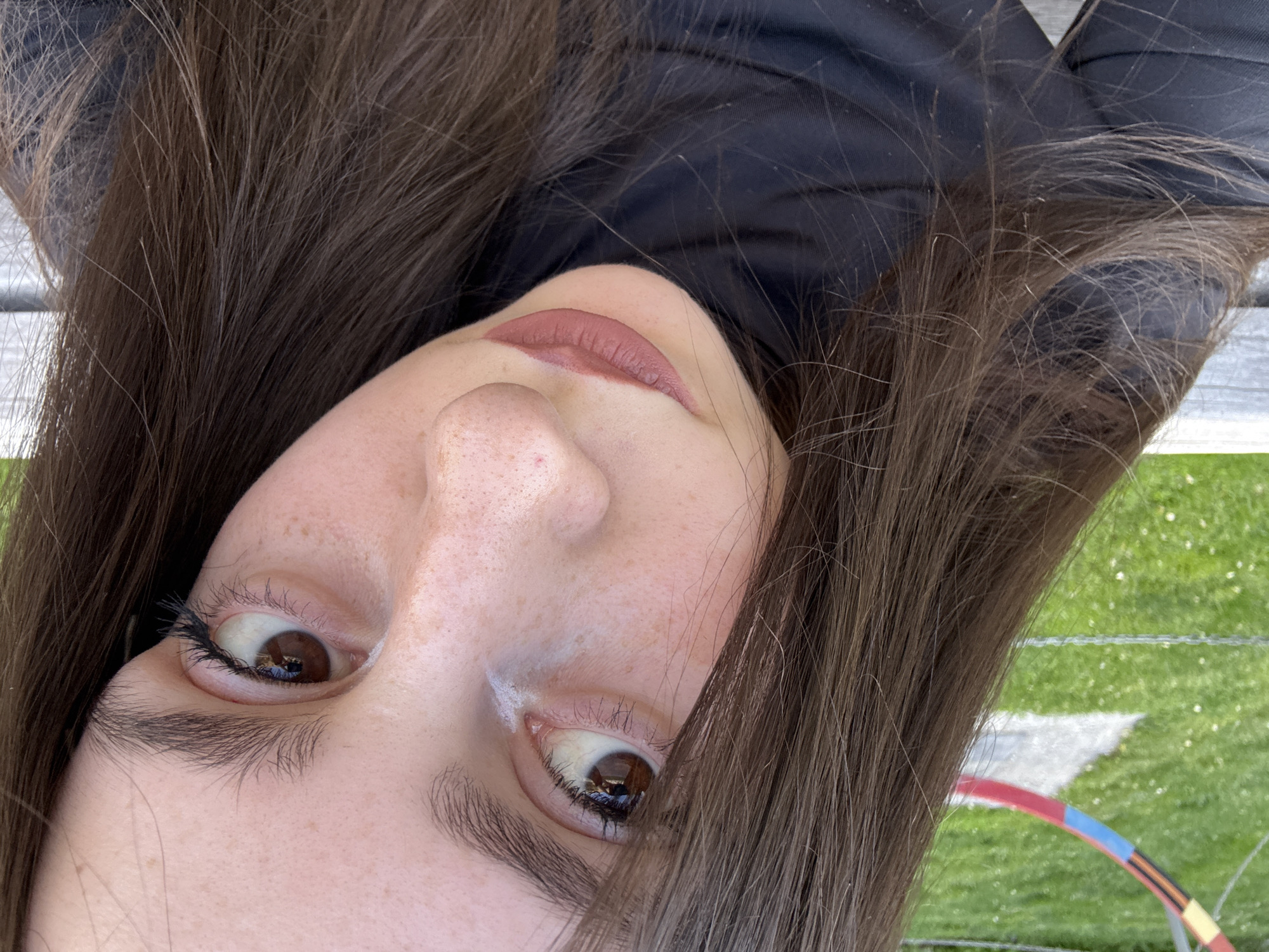 A close-up, upside-down portrait of a young woman with brown hair, brown eyes, and fair skin with freckles. She is wearing a dark-colored, possibly black, turtleneck. The background shows a hint of outdoor scenery, including green grass and what appears to be a colorful object, possibly a hula hoop or part of a play structure.
There is no verifiable evidence that can confirm the location in the city of Yerevan, Armenia.