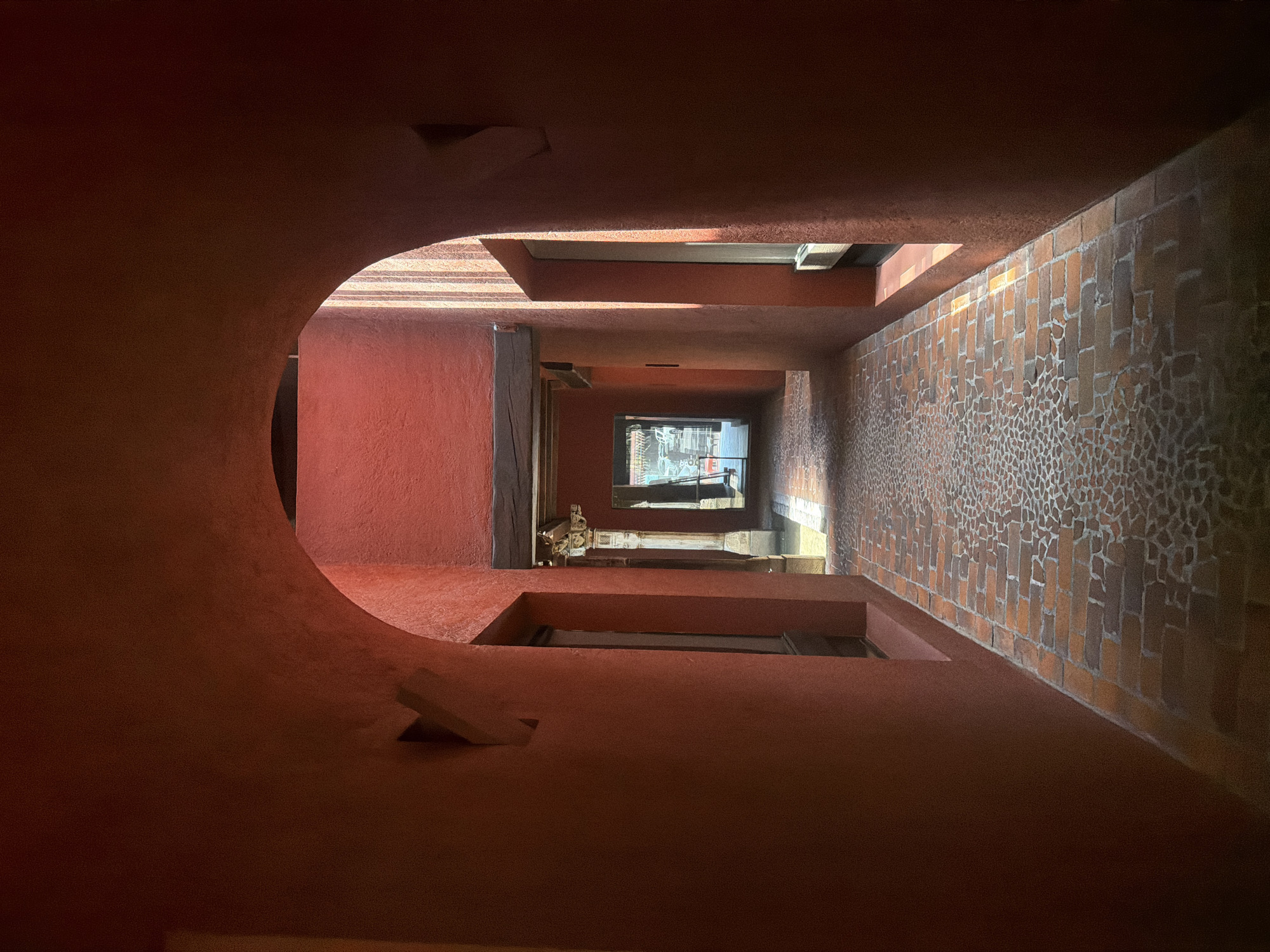 The image depicts a view from below, looking up through the interior of a building with red walls and ceiling. The walls have rounded cut-outs and rectangular insets, creating a layered effect. One wall features a brick pattern. In the distance, a window is visible, showing a glimpse of the outside world. The architecture appears to be in the style of Luis Barragán. It is located in Puebla, Mexico. There is no evidence of any people in the scene.
