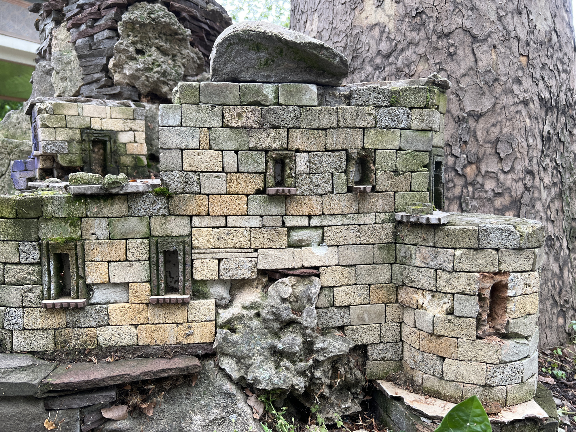 The image shows a miniature, weathered structure built of porous, tan and gray brick. The structure has architectural features, including small windows and crenellated walls. The structure appears to be part of a larger, more complex construction involving stacked stones and a large, tree trunk in the background.
The image does not provide any information about the city of Krasnodar, Russia, or anything to indicate its location. No people, subjects, actions, or interactions are visible.