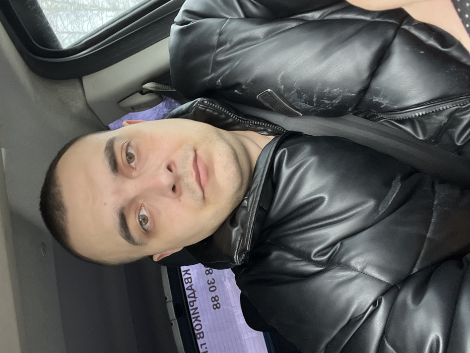 A male individual is depicted in a close-up, seated within the passenger compartment of a vehicle. He has short, dark hair, light-brown eyes, and a neutral facial expression, looking directly at the camera. He wears a black, padded, puffer-style jacket, secured by a dark seatbelt across his chest. The jacket exhibits scuff marks, particularly on the right frontal area. Through the rear window, reversed Cyrillic text and numbers, specifically "КВАРТИР 88 30 88" (APARTMENTS 88 30 88), are visible. Through the side window, bare tree branches are discernible against a light, overcast sky, indicating an outdoor environment. The vehicle's interior headliner appears light-colored. This scene is located in the city of Dubrovo, Russia.