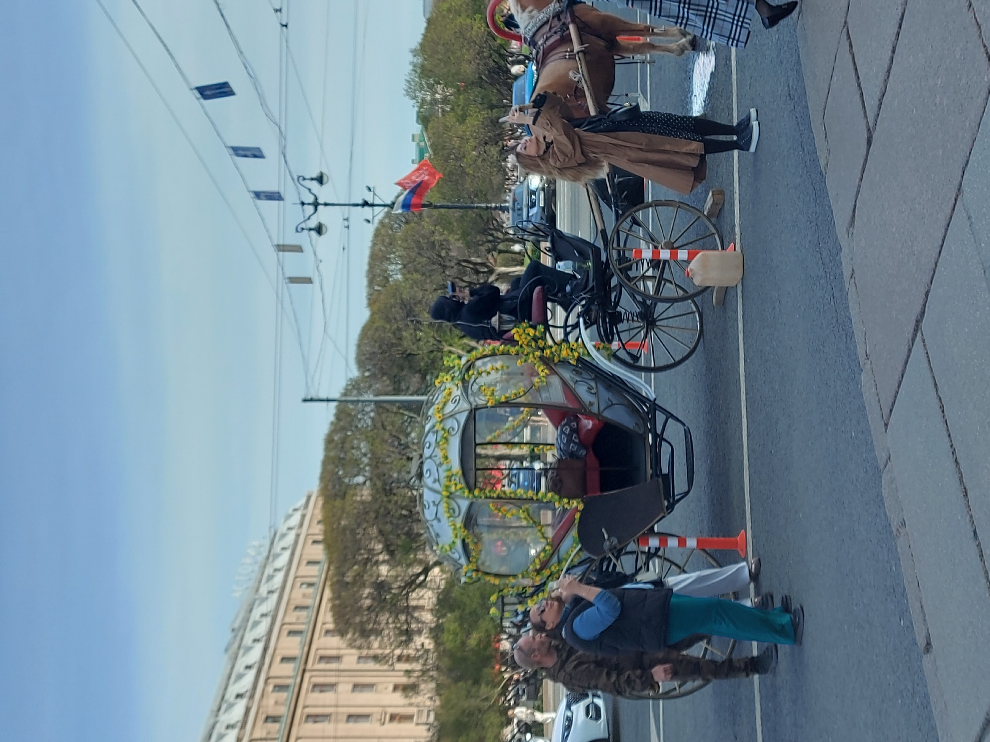 The image shows a scene in Saint Petersburg, Russia, with several horse-drawn carriages on a street, likely for tourist rides. At least three carriages are visible, with one decorated with yellow flowers around its glass frame. People are standing near the carriages, seemingly interacting with them or taking photos. A woman in a tan coat is taking a picture with her phone. The Russian flag is attached to a lamp post. A tall building stands in the background. The sky is clear and blue. Overhead wires and lamp posts run along the street. A horse is visible in the first carriage. The street is paved and has painted white lines separating lanes.