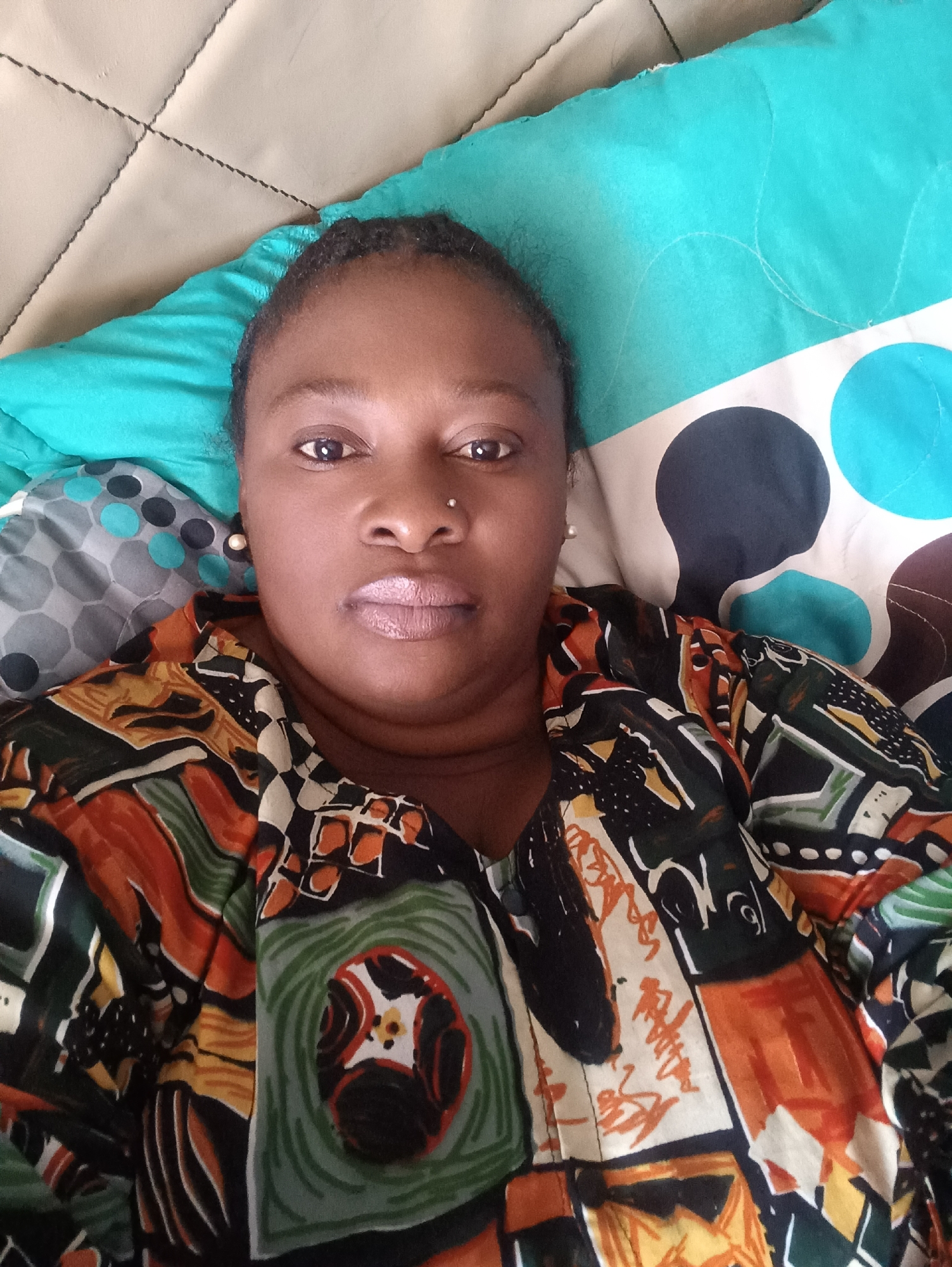 A woman is lying down on what appears to be a bed. She is wearing a colorful shirt with a geometric pattern, possibly made of orange, black, and green colors. She has dark skin, dark hair pulled back, and a nose piercing. The bed has pillows or cushions with different patterns, including polka dots and a quilted design. The image was likely taken indoors. The information provided does not confirm that this image was taken in Jos, Nigeria. No actions or interactions can be defined from the image.
