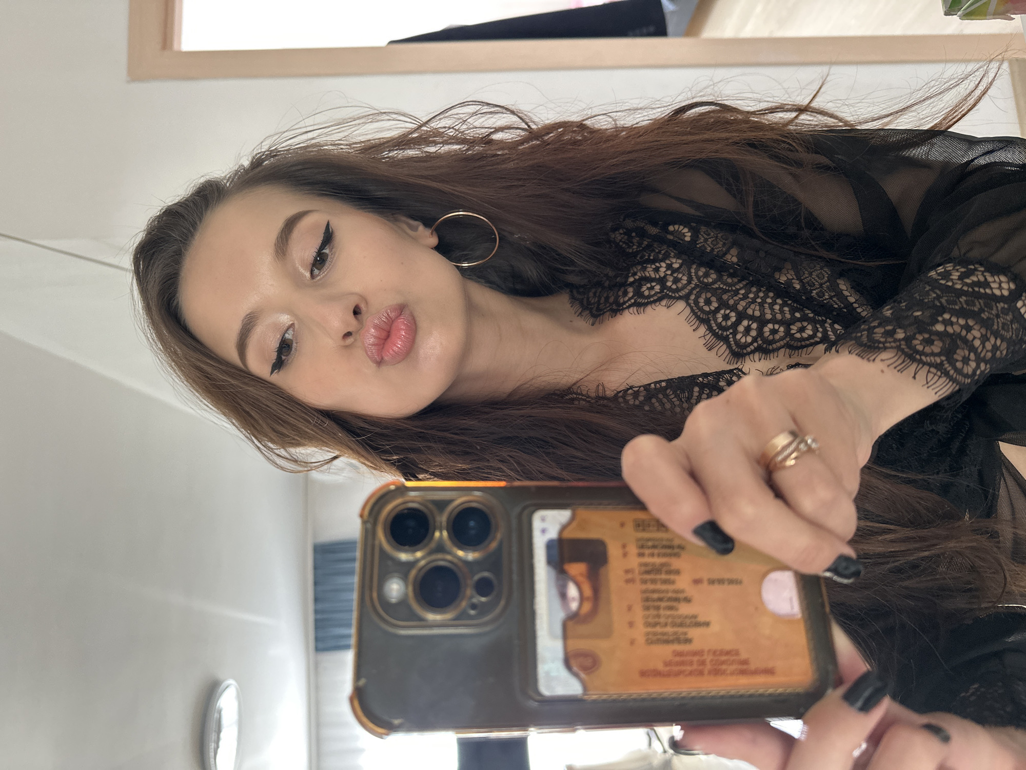 A young woman takes a mirror selfie in an indoor setting, likely a bathroom or bedroom, in Yekaterinburg, Russia. The image is oriented horizontally, showing the woman's upper body and face in a close-up shot. She has long, dark brown hair flowing around her shoulders and is wearing a black sheer top or robe with intricate lace detailing over what appears to be lingerie.
Her face is angled towards the camera, with her lips puckered in a "duck face" expression. She wears winged eyeliner, light eyeshadow, and lip gloss/lipstick. A gold hoop earring is visible on her right ear. On her right hand, which holds the smartphone, she wears multiple gold-toned rings, and her nails are painted black.
She is holding a smartphone, which appears to be an iPhone model with a multi-lens camera system, encased in a dark-colored and gold-accented protective cover. Partially visible inside the clear back of the phone case is an identification card or driver's license. Although much of the text on the card is blurry, discernible words and phrases include "LICENSED," "DRIVER," "NAME," "DATE OF BIRTH," and "ADDRESS," suggesting it's an official document. Some faint text like "SALAS FRANCE" is also visible.
The background is simple and bright, consisting mainly of a plain white wall reflected in the mirror. A light-colored wooden frame or trim is visible at the top, and in the reflection behind the phone, there's a hint of a blue striped object, possibly a towel or curtain. The overall impression is a casual, self-portrait taken in a personal space.