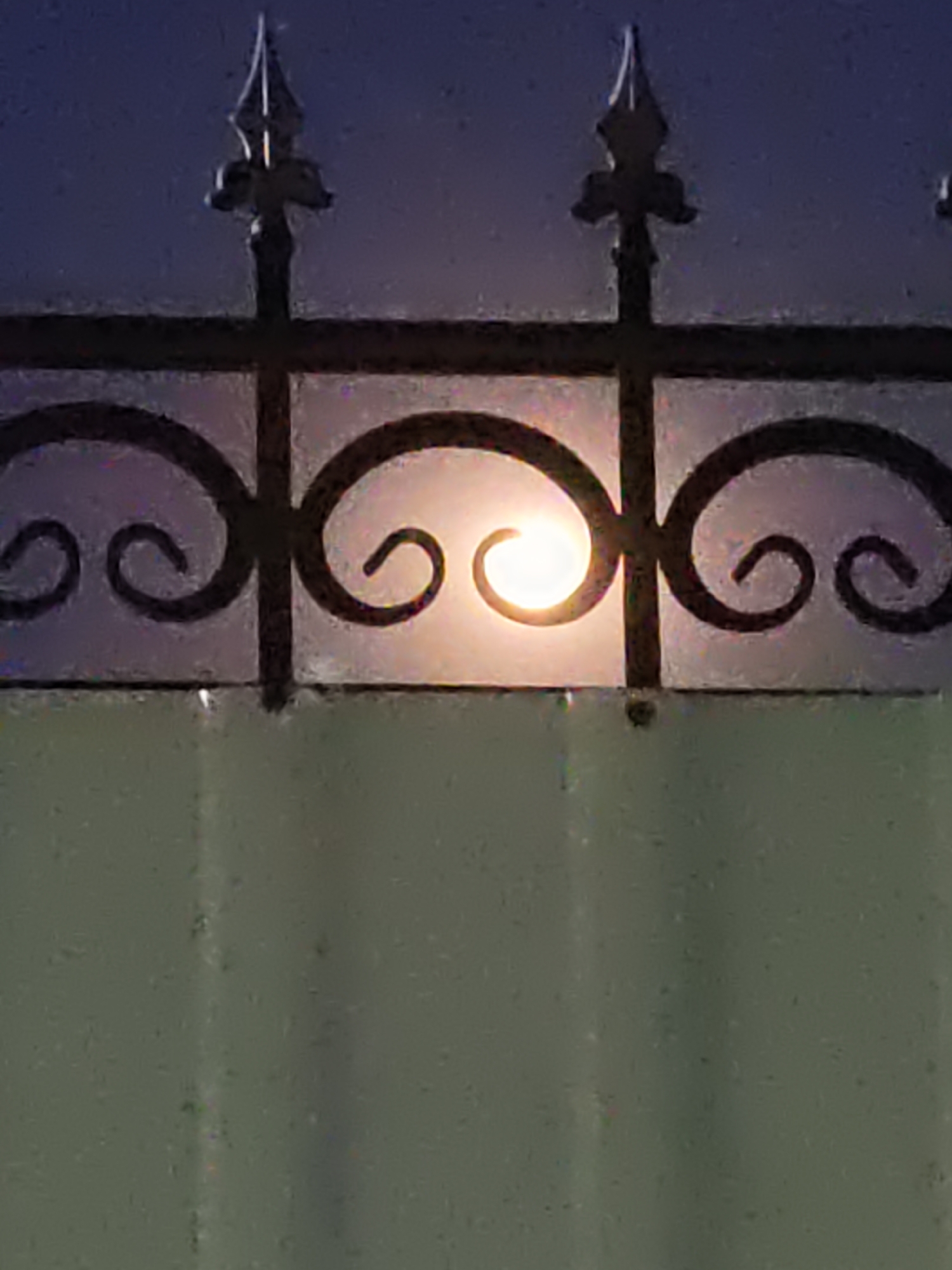 A nighttime image shows a black wrought iron fence in the foreground, with ornate scrollwork and pointed finials. Behind the fence, a bright, out-of-focus light source, likely the moon, is visible through a frosted or translucent panel. The background above the fence is a dark, hazy blue. Below the fence, a textured, pale green surface occupies the lower portion of the image. The scene appears to be outdoors, possibly a gate or a section of a perimeter fence. There are no people or discernible actions taking place. The location is identified as Rostov-on-Don, Russia.