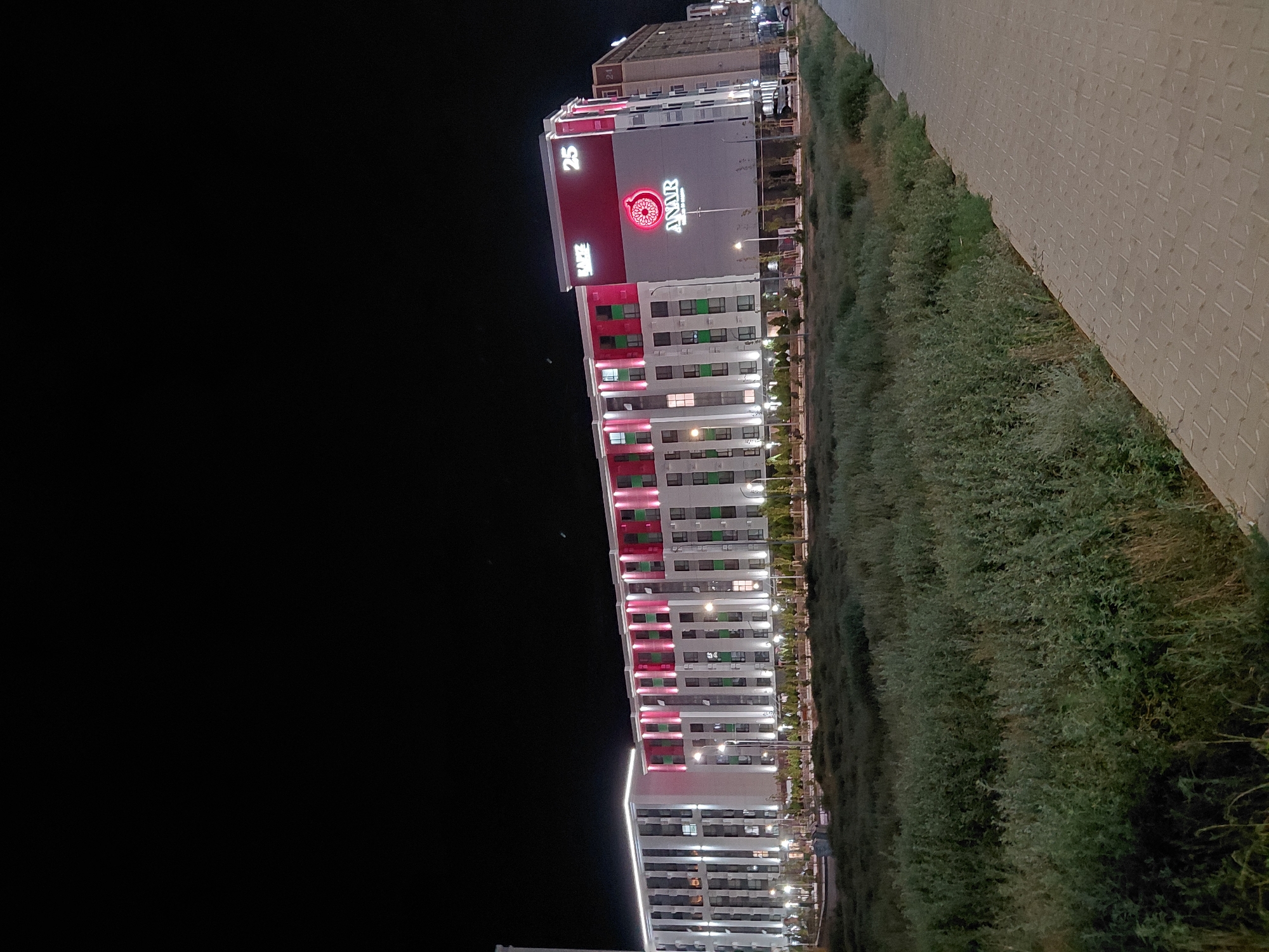 This is a nighttime exterior photograph of a modern, multi-story residential or commercial building complex in Aktau, Kazakhstan. The building is illuminated with a combination of white and pink lights, highlighting its architectural features, including rows of windows and decorative elements on the facade. A large, prominent red sign with a pomegranate logo and the word "ANAR" is visible on one section of the building, along with the number "25" and the word "RAFE" in white lettering. A paved walkway runs along the right side of the image, adjacent to a densely vegetated area that appears to be a park or undeveloped land. The sky is dark, indicating it is nighttime. There are no people or vehicles clearly visible in the immediate foreground or on the building itself.