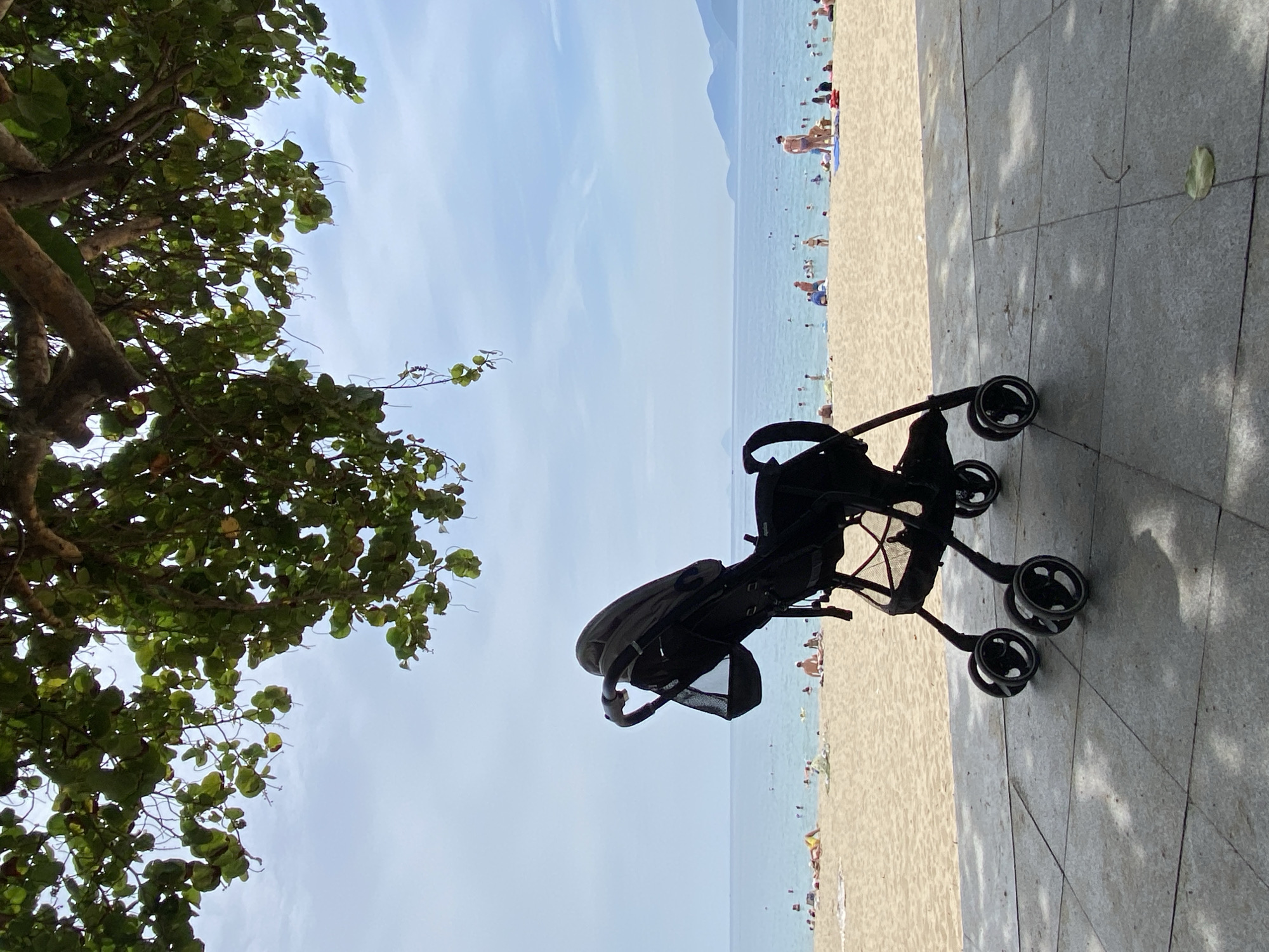 An empty, black baby stroller is parked on a paved walkway overlooking a crowded beach in Nha Trang, Vietnam. The stroller is in the foreground, positioned to the left of the frame. Beyond the walkway, a sandy beach is dotted with numerous people sunbathing and swimming in the calm, blue ocean. The water is clear, and in the distance, a hazy mountain range can be seen along the coastline. Overhead, the branches of a large tree with lush green leaves cast dappled shadows on the walkway, framing the scene. The sky is mostly clear with a few wispy clouds. The overall atmosphere suggests a warm, sunny day at a popular tourist destination.