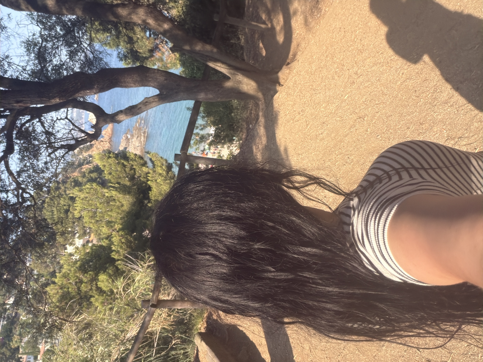 A first-person perspective selfie captures a person with long, dark hair from the shoulders up, standing on a dirt path overlooking the Mediterranean Sea. The person is wearing a black and white striped shirt. The background features lush green trees and bushes, a wooden fence, and the blue sea with rocky outcrops and a small beach in the distance. The scene is lit by bright sunlight, casting shadows on the ground and the person's shoulder. The location is the coastal path near the botanical gardens of Blanes, Spain.