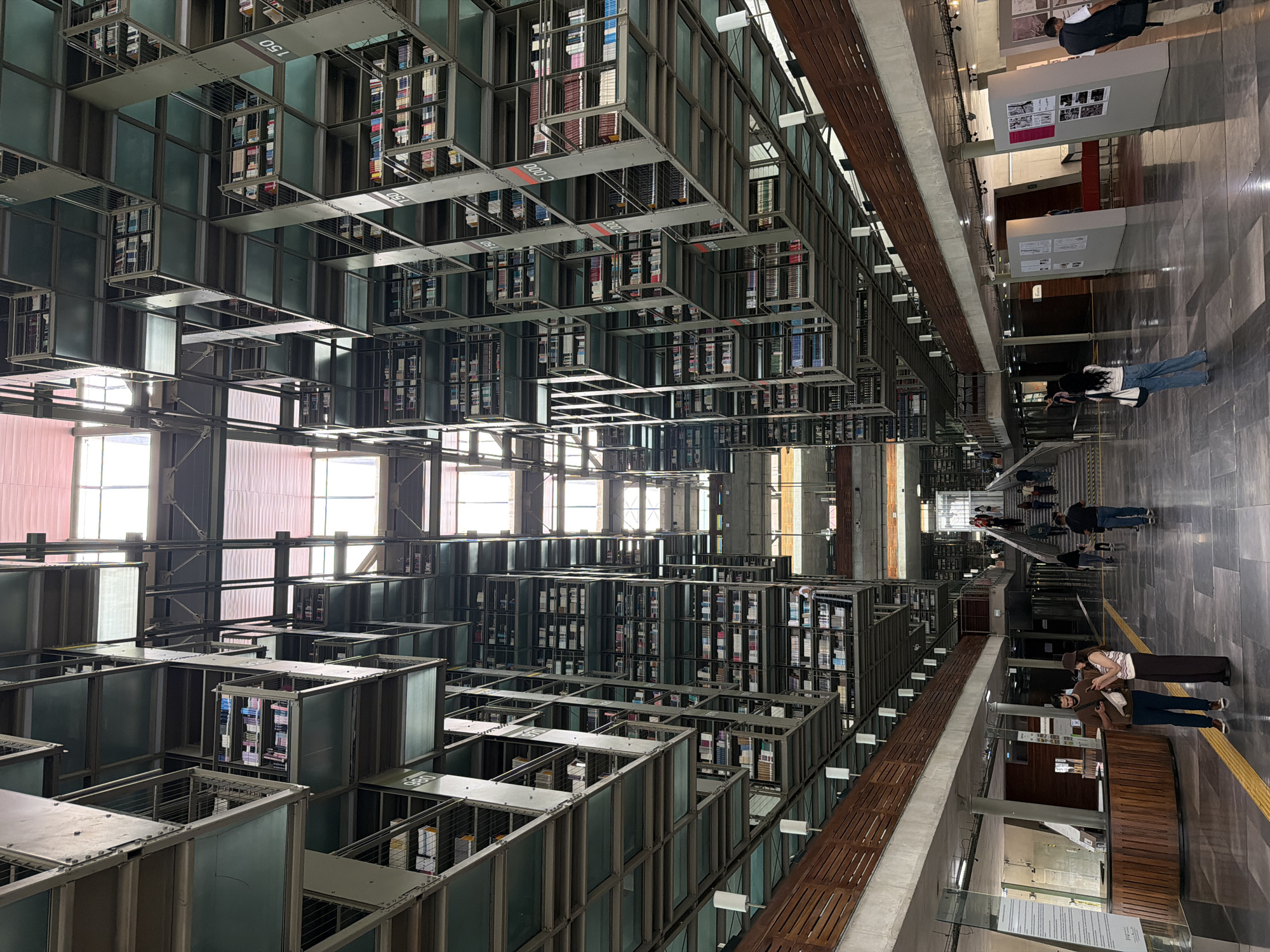 The photograph captures the interior of a modern library, characterized by its striking, multi-level shelving units filled with books. The architecture is industrial, with exposed metal structures and walkways suspended between towering bookshelves. Natural light filters in from a large, skylight-like opening at the top of the building, illuminating the vast space.
In the foreground, two individuals, a man and a woman, stand on a polished gray floor, both looking at their mobile phones. The man is wearing a dark t-shirt, blue jeans, and sneakers. The woman is wearing a striped top, wide-leg pants, and a hat. They are positioned near a wooden reception desk.
Further into the library, several other people are visible, some walking towards a large staircase in the distance, while others are scattered throughout the space. The upper levels of the library are densely packed with bookshelves, creating a sense of immense scale and architectural grandeur. Some shelves are labeled with numbers like "980," "570," "150," and "C 000."
The environment suggests a busy academic or public library. The presence of display boards with graphics on the right side of the image indicates an exhibition or informational area.
This library is the Central Library of the National Autonomous University of Mexico (UNAM), located in Mexico City, Mexico. The building's distinctive design is a well-known architectural landmark.
