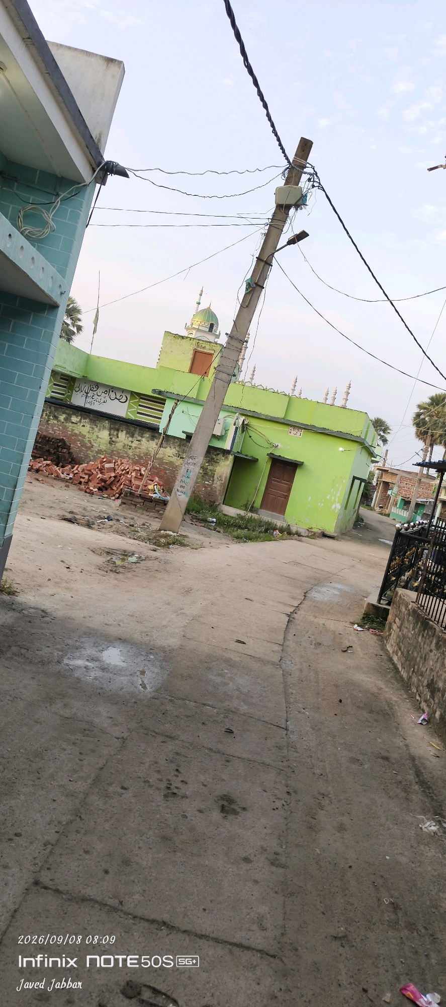 In Bahera, India, a street scene unfolds. A lime green building with a domed minaret stands as a central focus, likely a mosque. Bricks are piled on the ground in front of it, suggesting construction or repair work. A utility pole, laden with wires and a street lamp, leans precariously in the foreground, casting a shadow on the dirt and cracked asphalt road. The road itself is uneven, with some puddles of water visible. On the right, a black metal fence runs along a low wall, and a glimpse of other buildings and palm trees suggests a somewhat rural or semi-urban setting. The sky is overcast, hinting at a possibly warm day. The bottom right corner displays a watermark with a date and time, "2026/09/08 08:09," and a phone model, "Infinix NOTE 50S SG+," along with a name, "Javed Jabbar."