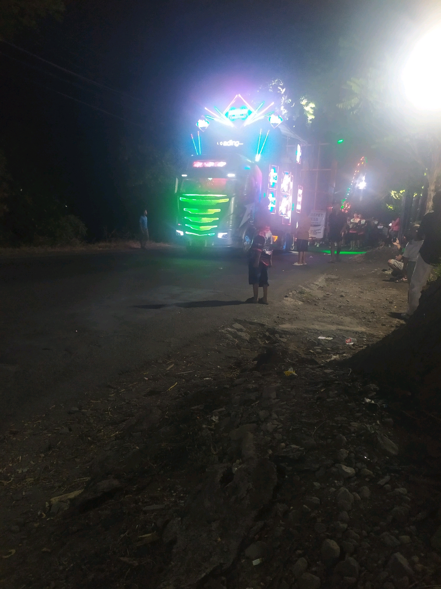 A nighttime exterior scene shows a heavily decorated truck with bright, colorful lights, likely used for a parade or event. The truck is parked on the side of a dark asphalt road, which appears to be in a rural or semi-rural area, with trees visible on the left and right sides of the road. The ground in the foreground is rough and uneven, with dirt, rocks, and some scattered debris. Several people are standing around the truck, some appearing to be spectators or participants in the event. One person, a young boy wearing a blue shirt and shorts, stands in the middle of the road, looking towards the truck. Another person, a young boy in a dark shirt and shorts, is standing to the right of the truck, facing away from the camera. The bright lights of the truck illuminate the area, creating a festive atmosphere. The overall scene suggests a lively nighttime gathering centered around the illuminated truck. The location is identified as Winong, Indonesia.