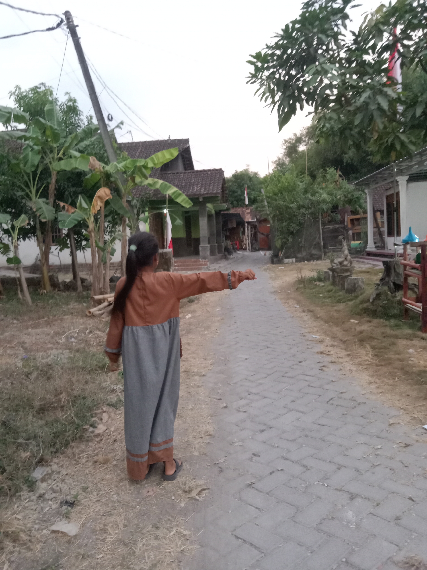 The image depicts an outdoor scene in a village setting, likely in Jumput, Indonesia, during daylight hours. In the foreground, a young person, seen from the back, is walking away from the camera down a cobblestone path. The person is wearing a long, brown and grey garment and dark sandals. Their right arm is extended outwards. The path is flanked by dry grass and dirt. To the left of the path are banana trees and some structures resembling houses with tiled roofs. To the right of the path, there is more vegetation, a white building with columns, and a wooden fence. In the background, there are more trees and houses, suggesting a rural or semi-rural residential area. Several utility poles and wires are visible against the overcast sky.