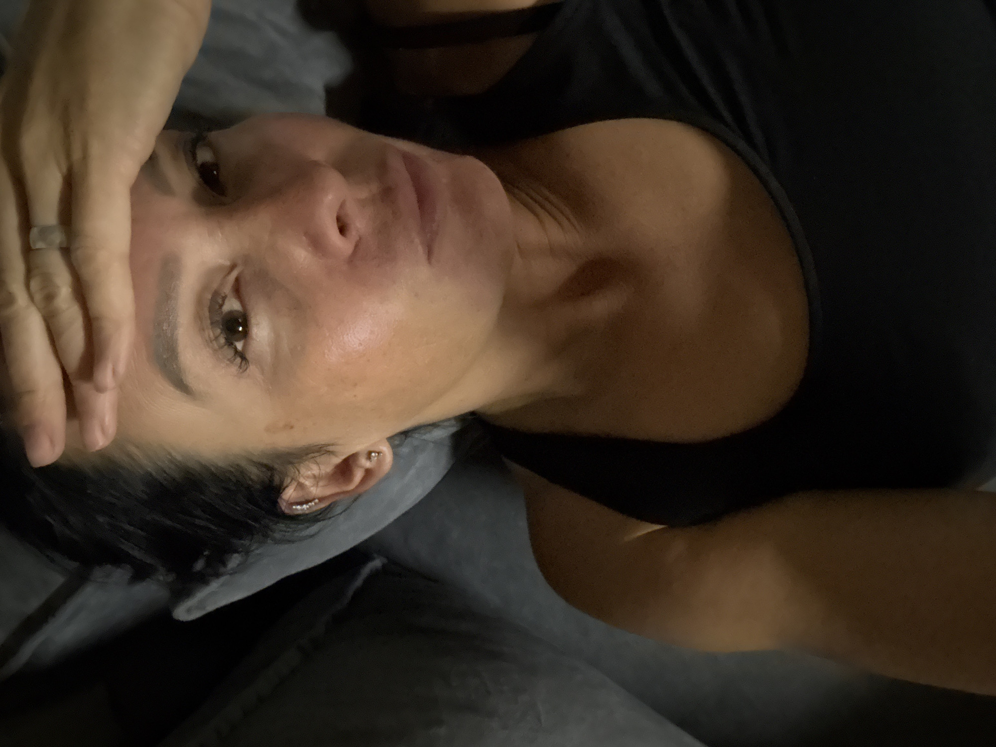 The image shows a close-up, overhead shot of a woman lying down. She has dark hair pulled back, and her right hand is placed on her forehead. She is wearing a black tank top. She has multiple earrings in her right ear, including a cuff and a stud. A ring is visible on her right ring finger. The background appears to be a dark, textured surface, possibly a couch or bed. There are no visible elements that indicate the location is in the city of Chaikovsky, Russia.