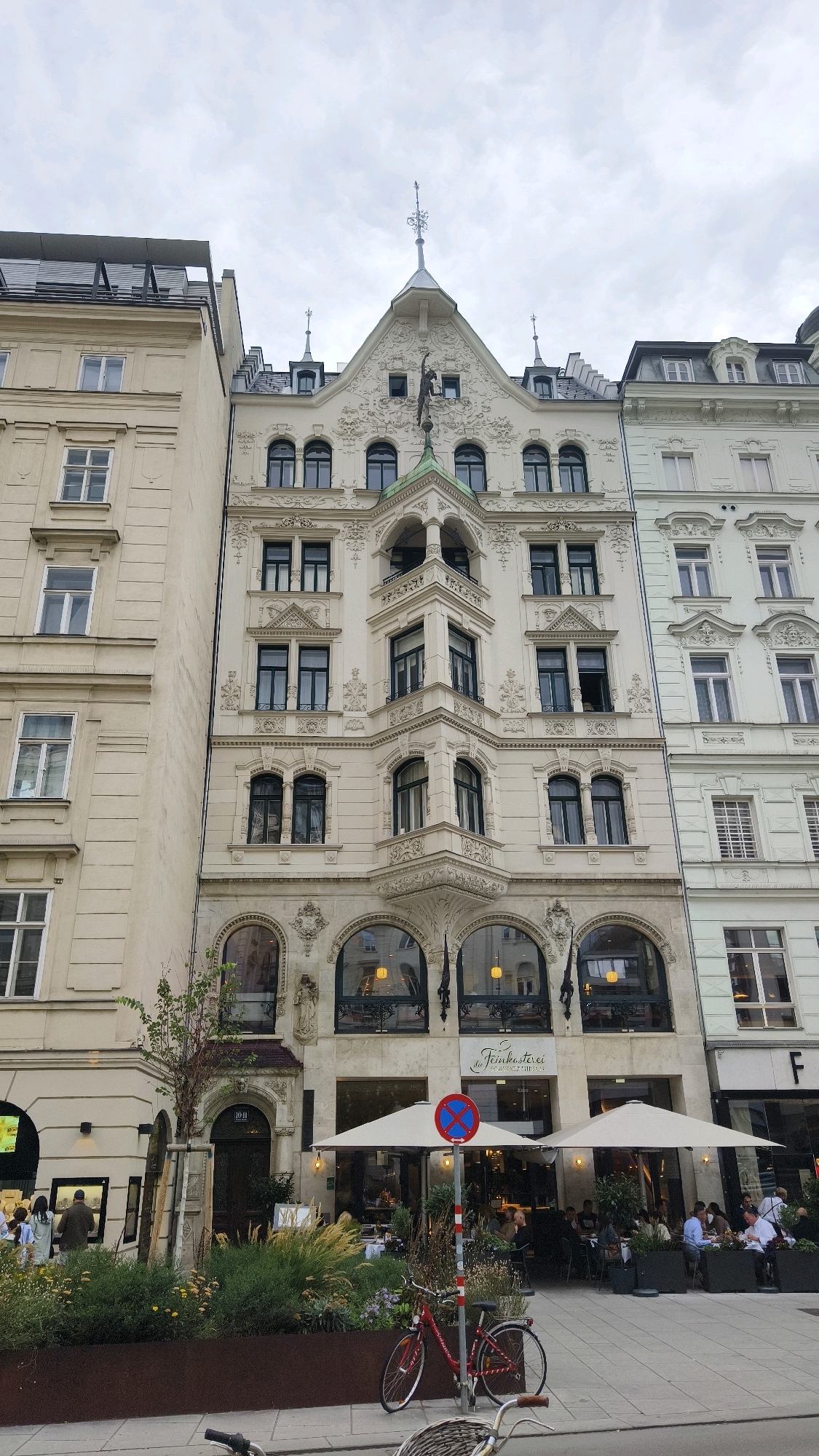 The image depicts a street scene in Vienna, Austria, featuring a row of ornate buildings and an outdoor cafe. The central building is cream-colored with intricate white stucco detailing, featuring a prominent triangular gable topped with a spire. It has multiple stories with numerous windows, some arched and some rectangular. The ground floor of this building houses a cafe named "de Feinkosterei." Two large cream-colored umbrellas shade tables outside the cafe, where several patrons are seated. A red bicycle is parked on the sidewalk in front of the cafe, and a red and blue no-parking sign is visible. To the left of the central building is a lighter yellow building with a simpler facade and rectangular windows. To the right is a pale green building with similar architectural detailing to the central one. The sky is overcast with clouds. The overall impression is a charming European street with a lively cafe atmosphere.