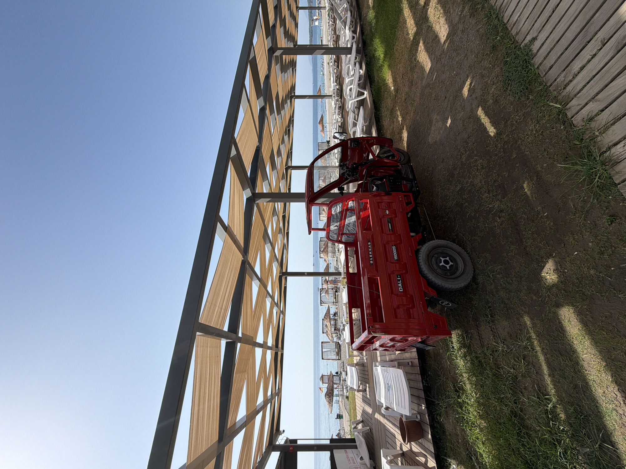The image depicts a bright, sunny day at a beach resort in Kemer, Turkey. A red three-wheeled vehicle, possibly a utility cart or a small truck, is parked on a dirt path next to a wooden deck. The vehicle has the branding "DALI- 15000S" visible on its side. In the background, a beach lined with lounge chairs and umbrellas stretches towards the turquoise sea under a clear blue sky. A covered walkway with a tan fabric canopy provides shade over the deck area. The environment suggests a tourist destination with amenities for relaxation and beach access.