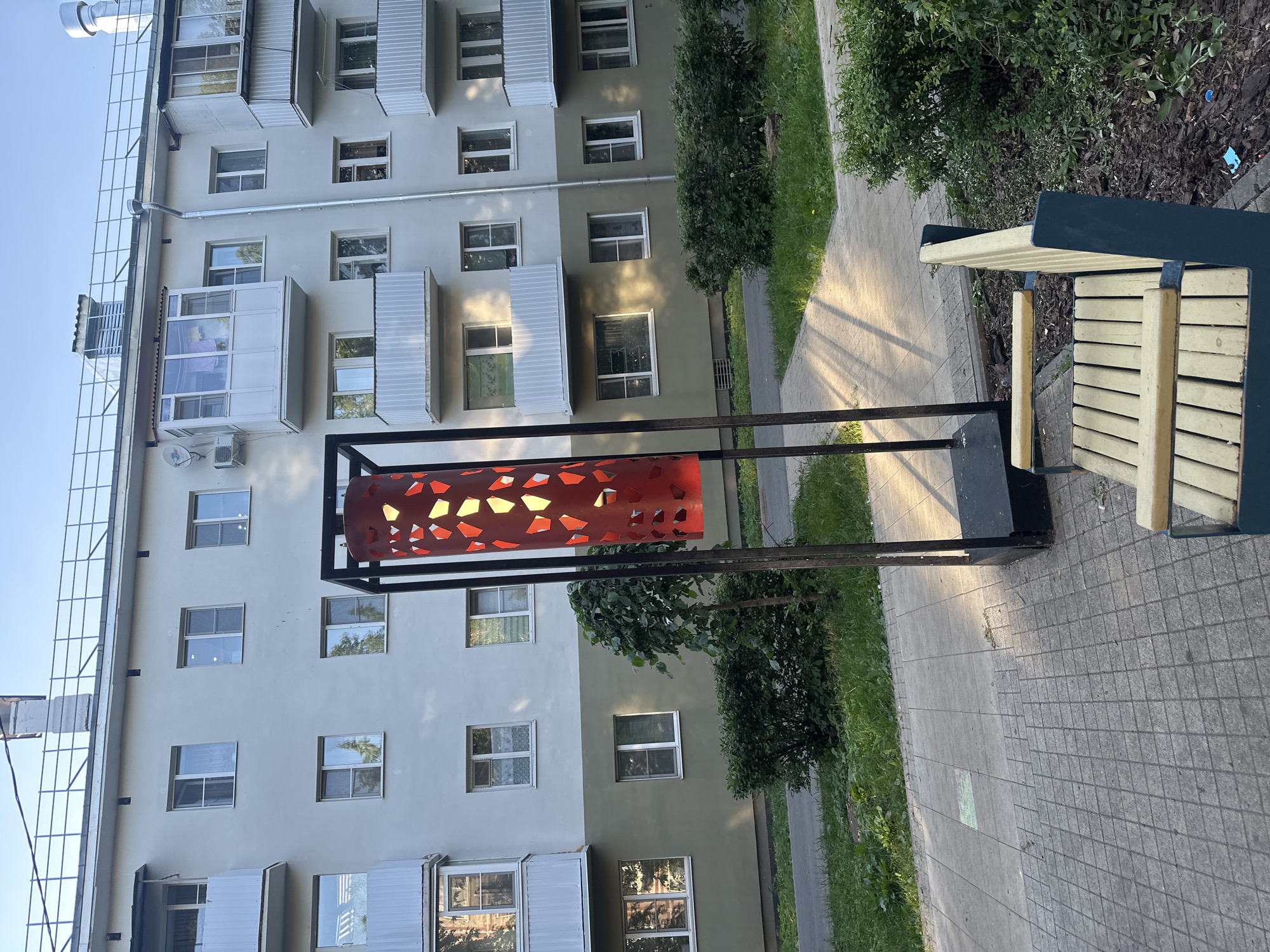 The image depicts a residential building in Vologda, Russia. In the foreground, there is a bench and a decorative lamp post with a red, patterned shade. The building, which appears to be multi-story, has numerous windows and balconies. The area surrounding the building includes a paved walkway, green grass, and bushes. The lighting suggests it is daytime, with sunlight casting shadows on the ground. There are no people visible in the image.