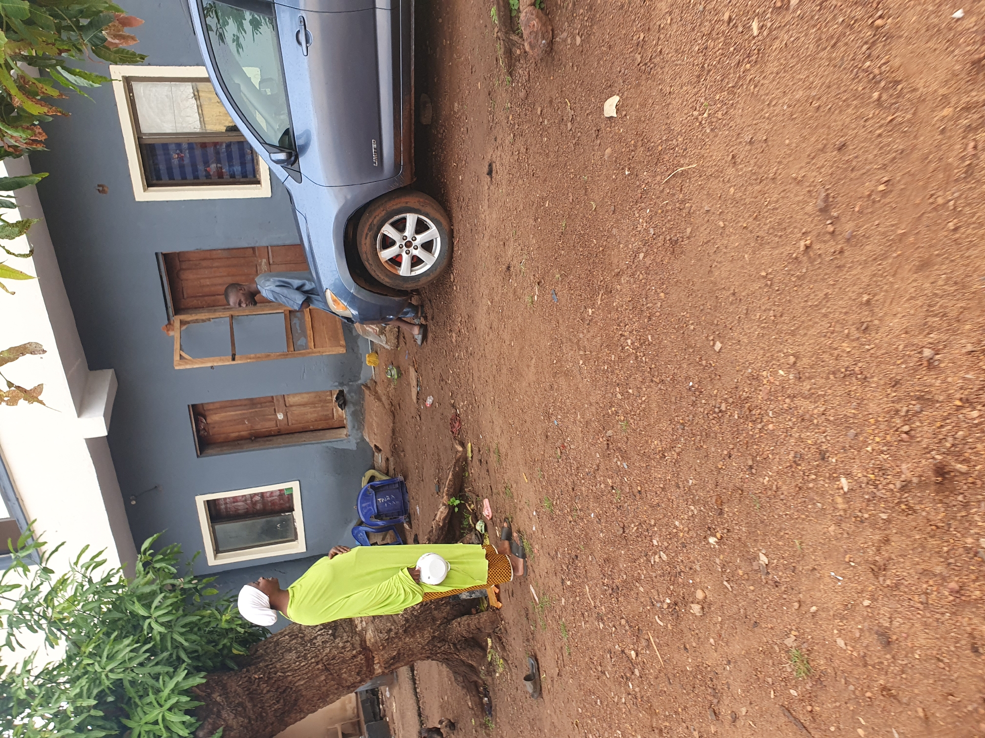 The image depicts an outdoor daytime scene in Idah, Nigeria, featuring two individuals, a car, and a building on an unpaved, reddish-brown dirt and gravel ground.
On the left, a man is seen standing next to a blue SUV, which appears to be a Toyota RAV4 with the word "LIMITED" visible above its rear wheel. He is wearing a light-colored, long-sleeved top and dark trousers. The man's face is visible, and he is looking towards the left.
To the right, a woman is captured in a lime-green long dress and a white headscarf. She is holding a small white bowl or container in her hands and facing towards the right side of the frame. Nearby, a blue plastic chair is present, bearing the word "TARA" on its backrest.
A large tree with green leaves, possibly a mango tree, stands prominently in the foreground on the left. In the background, a building with dark grey walls is visible, featuring several wooden-framed windows and wooden doors. The bright lighting suggests it is daytime with fair weather.