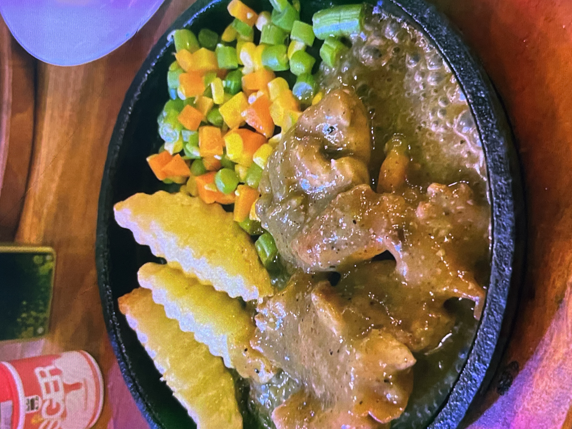 This is a close-up, overhead shot of a sizzling steak dish served on a black cast-iron platter, placed on a wooden surface. The platter contains pieces of steak in a rich, brown gravy, accompanied by a medley of diced vegetables, including green beans, carrots, and yellow bell peppers. Three golden-brown potato wedges with ruffled edges are arranged on the side.
In the bottom left corner, a partially visible red and white cylindrical object, possibly a can or a small container, bears some text in what appears to be an Asian script, with the characters "F" and "T" discernable. To the left of this object, a small green rectangular item, possibly a phone or a coaster, is also visible.
The overall scene suggests a meal being served, likely at a restaurant given the presentation on a sizzling platter and the presence of accompanying items. The lighting suggests an indoor setting, possibly with artificial light, as there are no clear indications of time of day or weather. The image is primarily focused on the food, highlighting its texture and colors. The focus is sharp on the food, with a slight blur in the background, emphasizing the culinary subject. No people are visible in the frame. The cuisine appears to be a Western-influenced dish, possibly adapted to Indonesian tastes, given the provided location context of Cadek, Indonesia.