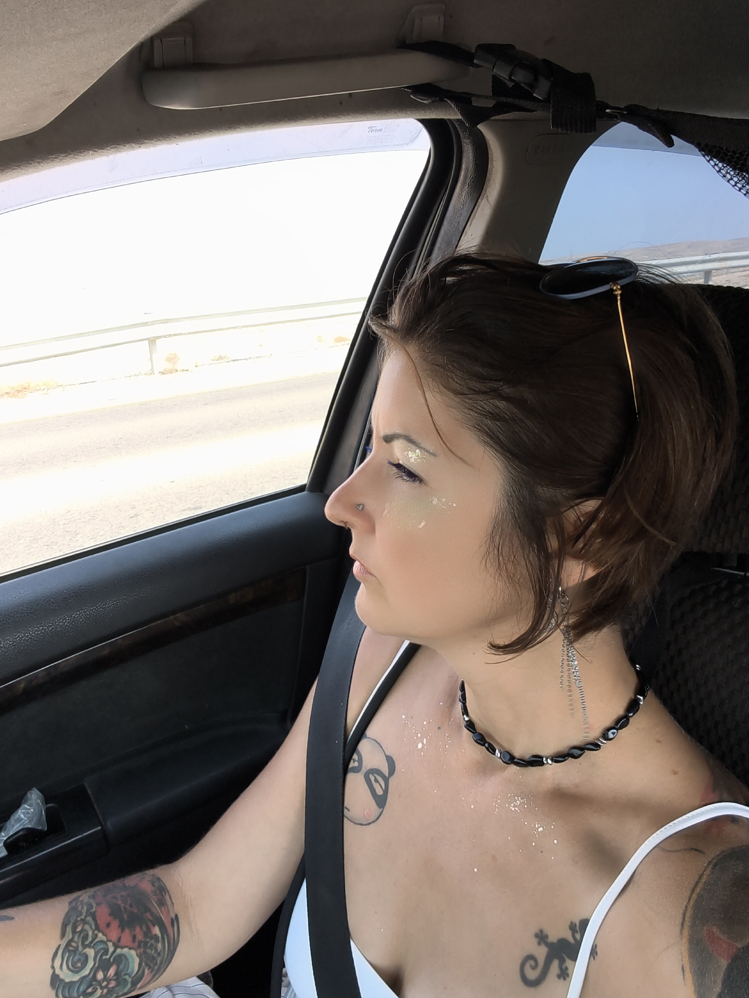 A woman with short brown hair and glitter on her face is sitting in the passenger seat of a car. She is wearing a white tank top, a black beaded necklace, and a seatbelt. Her arm is visible, covered in tattoos. The car appears to be driving on a road, with a blurry landscape visible through the window. The sun is shining brightly. The location is the Ramat Negev Regional Council in Israel.