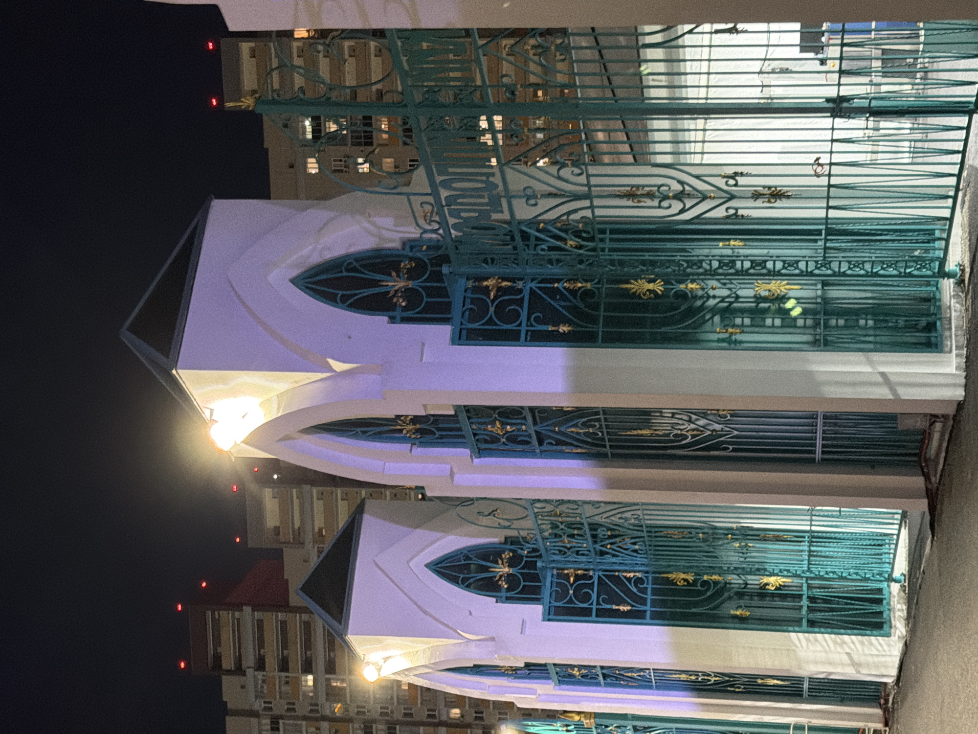 The photograph depicts a nighttime scene of an exterior walkway or street in front of a building with ornate, teal-colored metal gates and white architectural elements. The gates feature intricate scrollwork and gold accents. Behind the gates, a white wall with arched window-like structures is visible, illuminated with a purple light. The buildings in the background are tall, apartment-style structures with visible windows, some lit from within, and red lights at their apexes, indicating they are in a city environment. The overall impression is one of a formal entrance or facade, possibly of a religious or public building, at night.