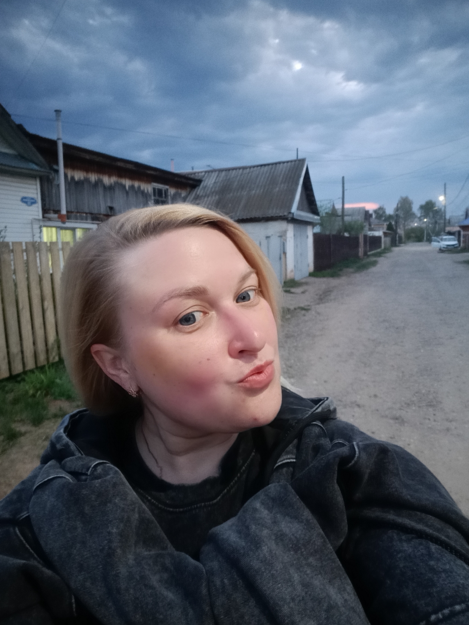A young woman is taking a selfie on a dirt road in what appears to be a residential area. She has fair skin, light blonde hair, and blue eyes. Her expression is playful, with pursed lips. She is wearing a dark-colored jacket or hoodie.
The background shows a typical Russian village street with wooden houses, fences, and a dirt road. The sky is overcast with dark clouds, suggesting it is either early morning or evening. In the distance, a car is parked on the side of the road. The scene appears to be in a rural or semi-rural area, possibly on the outskirts of the city of Chaikovsky, Russia.