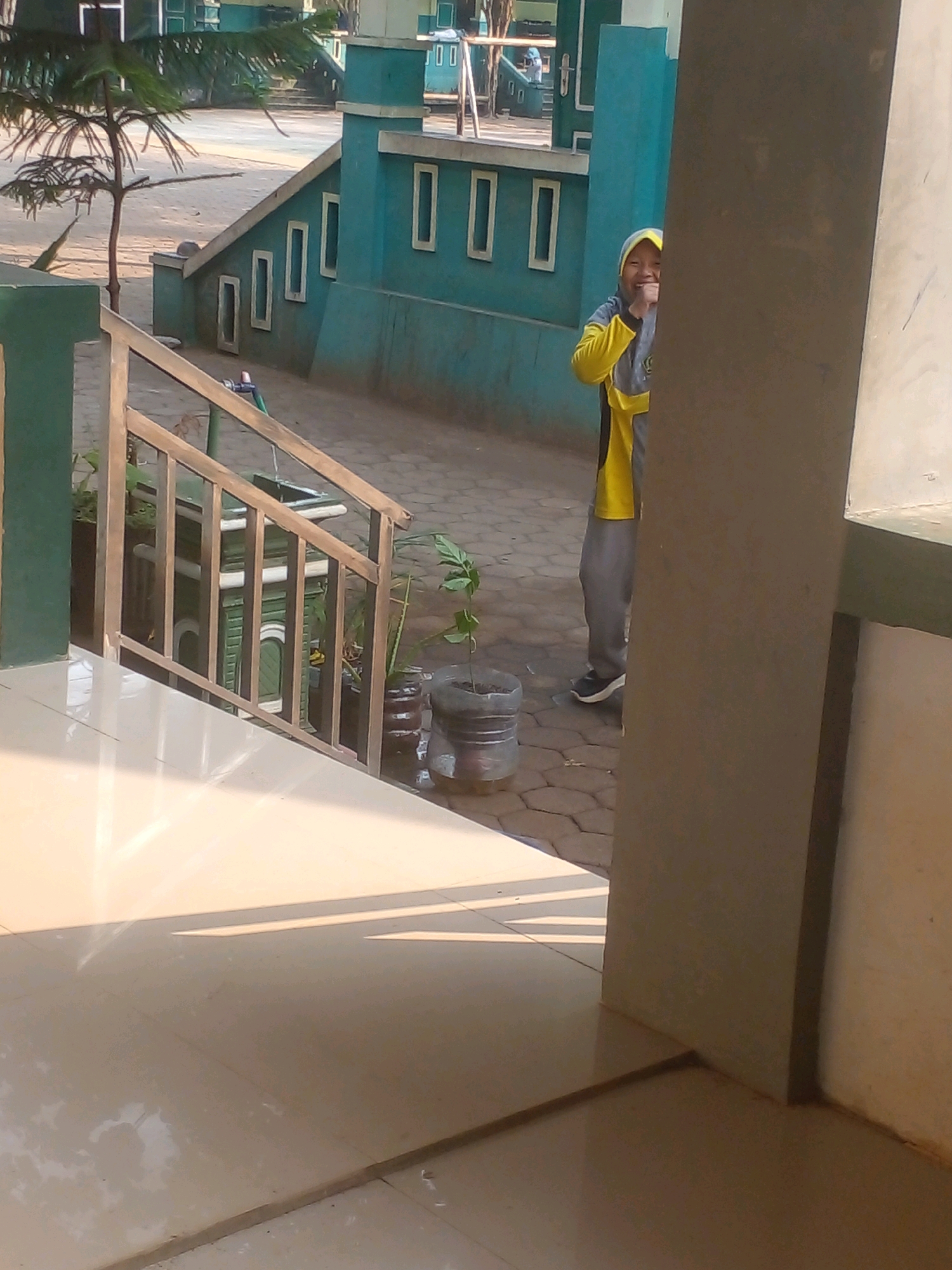 A young person wearing a yellow and grey hoodie and grey pants stands on a paved walkway, partially hidden behind a thick pillar. The person is smiling and has one hand raised to their face. The background shows a teal-colored building with white rectangular window frames, a metal railing, and some potted plants. The ground is paved with hexagonal tiles. The scene appears to be outdoors, possibly at a school or public facility, given the architectural style and the presence of a playground-like structure in the distance. The lighting suggests it is daytime, with shadows cast on the tiled floor. The location is identified as Banyuputih, Indonesia.