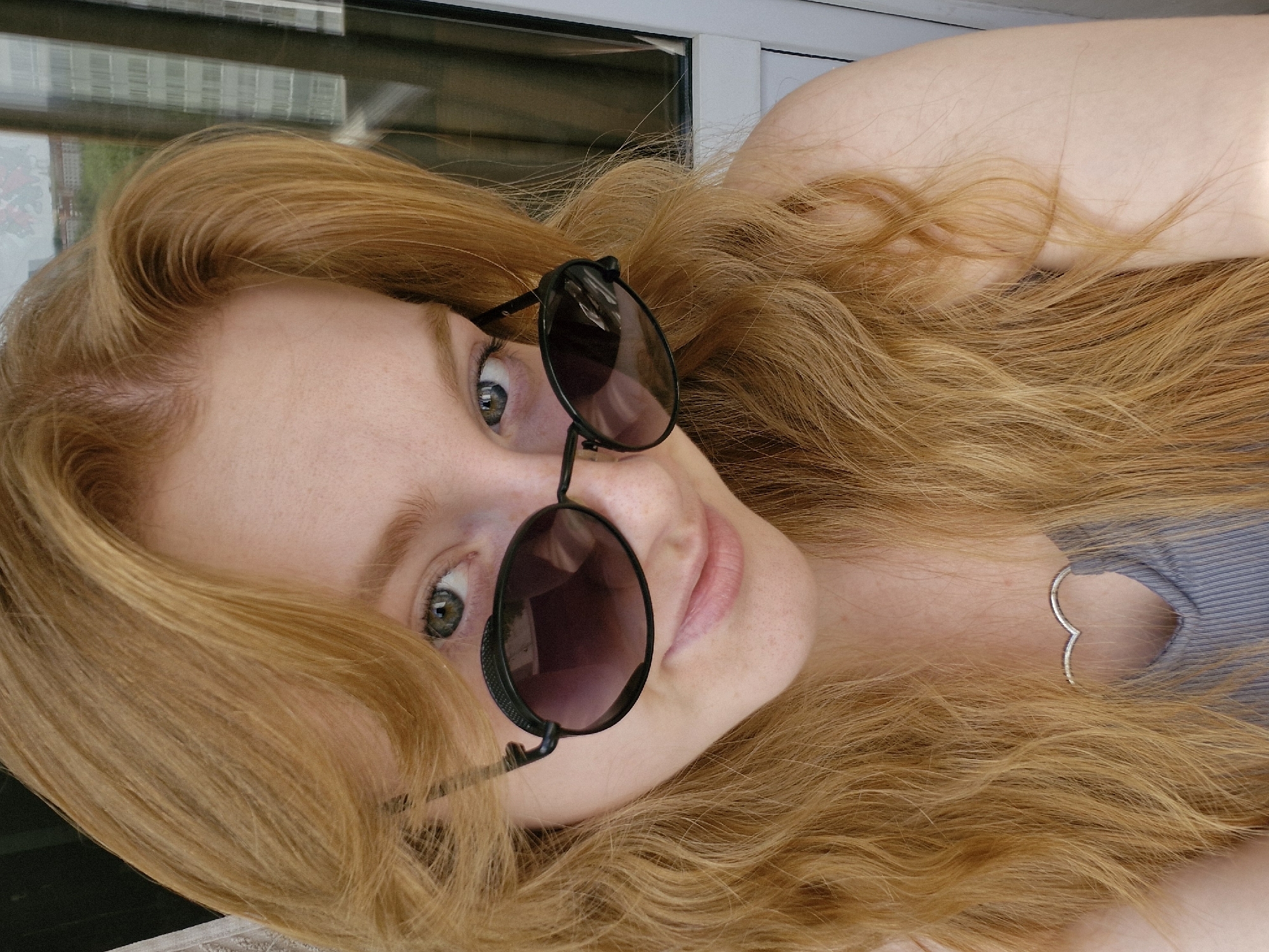 A light-skinned woman with wavy, strawberry-blonde hair is posing with sunglasses on her face. She is wearing a light-colored, ribbed tank top with a heart-shaped cut-out. Her skin is fair and her eyes are a light green. There is a window behind her that reflects a building. The location cannot be confirmed.