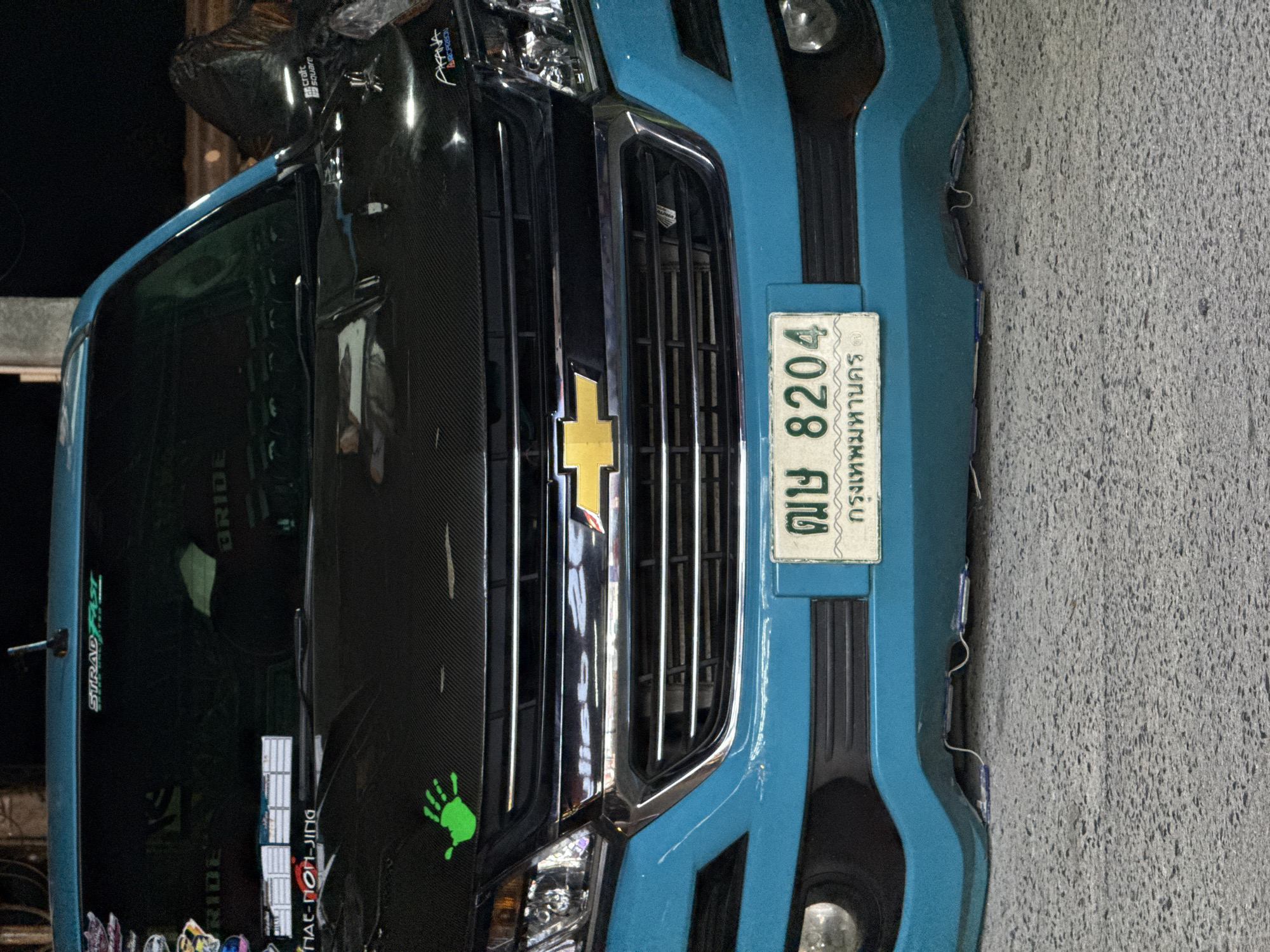 This is a close-up, low-angle shot of the front of a bright blue vehicle, possibly a truck or SUV, parked on a textured gray surface, suggesting a road or parking lot. The vehicle has a prominent chrome grille with vertical slats and a yellow Chevrolet emblem. The hood appears to be made of carbon fiber, adding a sporty detail.
A license plate is clearly visible on the right side of the front bumper. It is white with black text and numbers, and has Thai characters. The text reads "ฒษ 8204 กรุงเทพมหานคร" which translates to "GZ 8204 Bangkok".
There are various stickers and decals on the vehicle. On the left side of the windshield, partially obscured, are stickers with "STRADFAST" and "BRIDE". A green handprint sticker is visible on the carbon fiber hood, to the left of the grille. On the upper left of the vehicle, there is a sticker that says "crafc square".
The lighting suggests it is either nighttime or dusk, as the background is dark, and artificial lights are reflecting off the vehicle's surfaces. The overall impression is of a customized vehicle in a public space in Baan Bo Phut, Thailand, likely during an evening or night. There are no people visible in the frame.