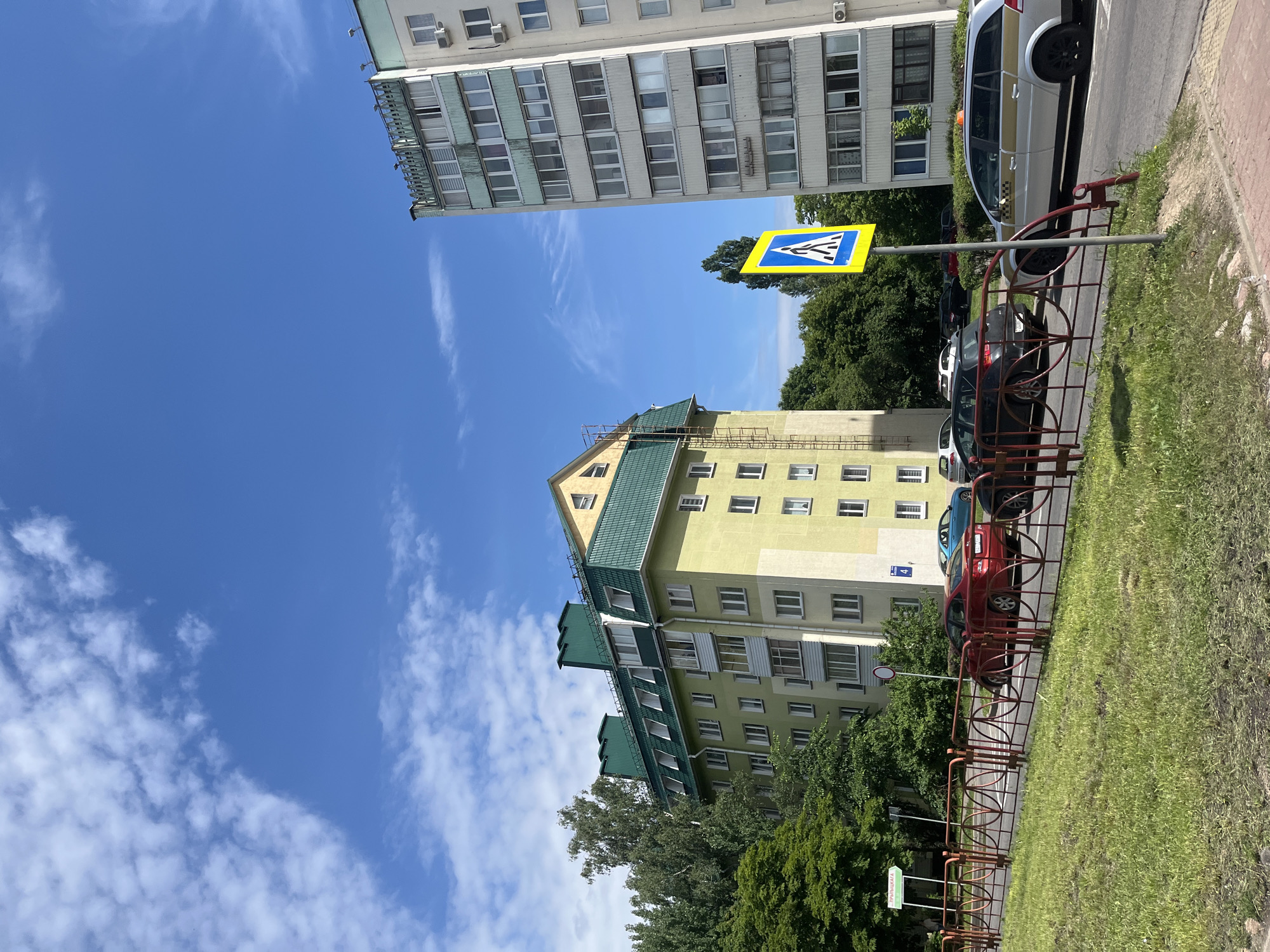 This is a daytime photograph capturing an urban scene in Minsk, Belarus, specifically at Pervomayskaya Street (Першамайская вуліца), near house number 4. The image is taken from a slightly low angle, looking towards a road lined with buildings, under a clear blue sky dotted with scattered white cumulus and wispy cirrus clouds.
In the foreground, a strip of green grass borders a low, decorative red metal fence with interconnected circular patterns. Beyond the fence is a paved road, along which several vehicles are parked. From left to right, these include a red sedan, a dark grey or black hatchback/SUV, and a silver station wagon with a gold stripe and an orange roof light, indicating it is a taxi. Further down the road, a light blue car is also visible.
Two prominent multi-story apartment buildings dominate the background. The building on the left features a light yellow-green facade and a distinctive dark green pitched roof with several dormer windows and angular green ventilation structures. A metal ladder extends vertically up the right side of this building. A blue house number plaque displaying "4" is affixed to its facade. Lush green trees and foliage are visible around and between the buildings, with some taller trees rising above the yellow-green building's roofline. Behind the red fence and in front of the yellow-green building, a green sign with white Cyrillic text reading "ПЕРШАЦКАГА" is partially obscured by trees.
To the right, a significantly taller, slender apartment building stands with a light grey or off-white facade. It features numerous rectangular windows and a flat roofline with decorative elements or railings. Several white air conditioning units are mounted on its exterior walls. A bright yellow pedestrian crossing sign, a square with a blue border and a white pedestrian icon, is prominently positioned in the middle ground, on the right side of the road.