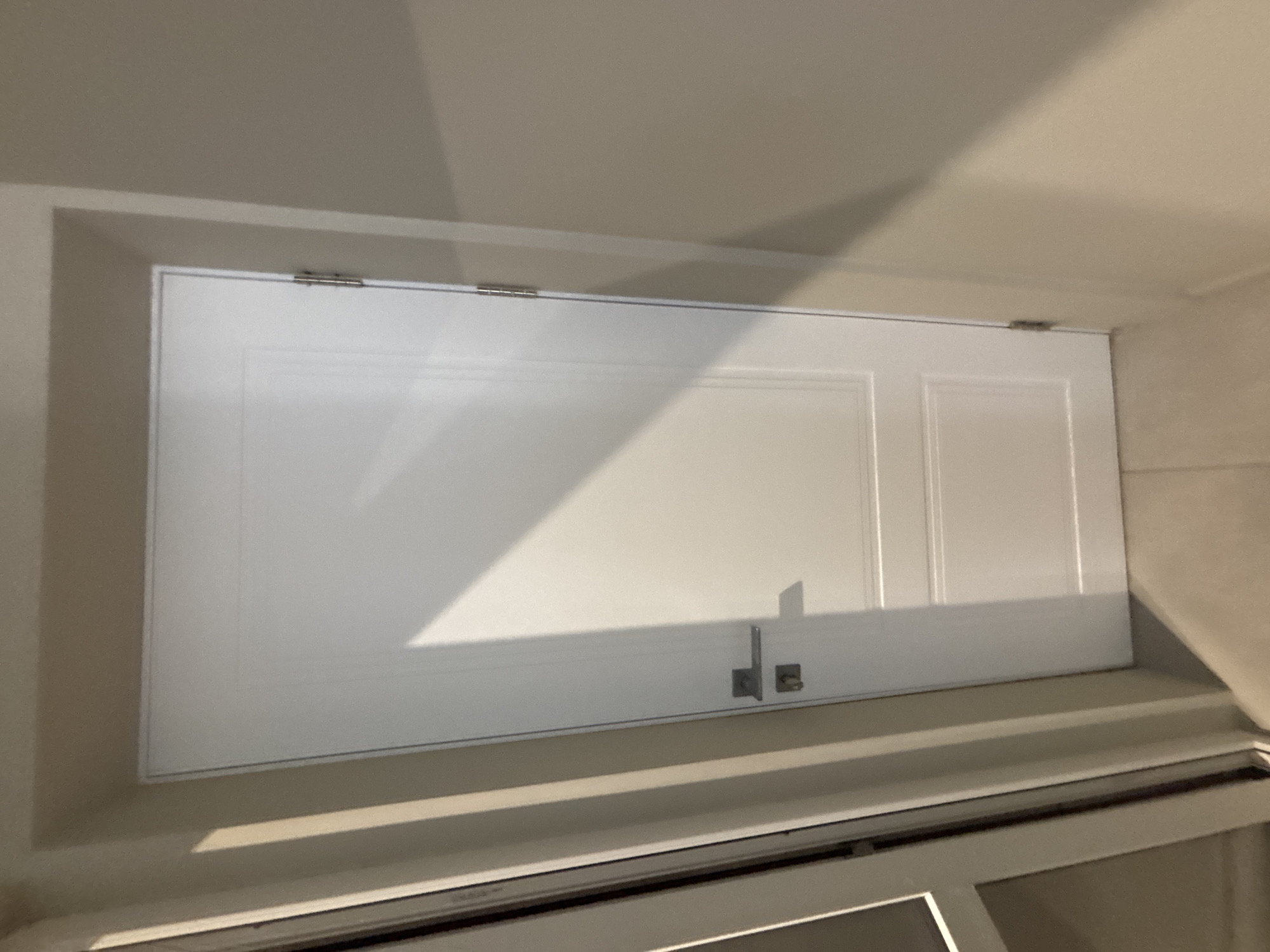 The image shows a white interior door with a silver doorknob and hinges, set within a beige wall. A portion of a glass-paned door or window frame is visible on the left edge of the image. Sunlight casts a diagonal shadow across the white door. The scene appears to be indoors, with the floor showing beige tiles. There are no people or animals visible. The location is Ldzaa, Abkhazia.