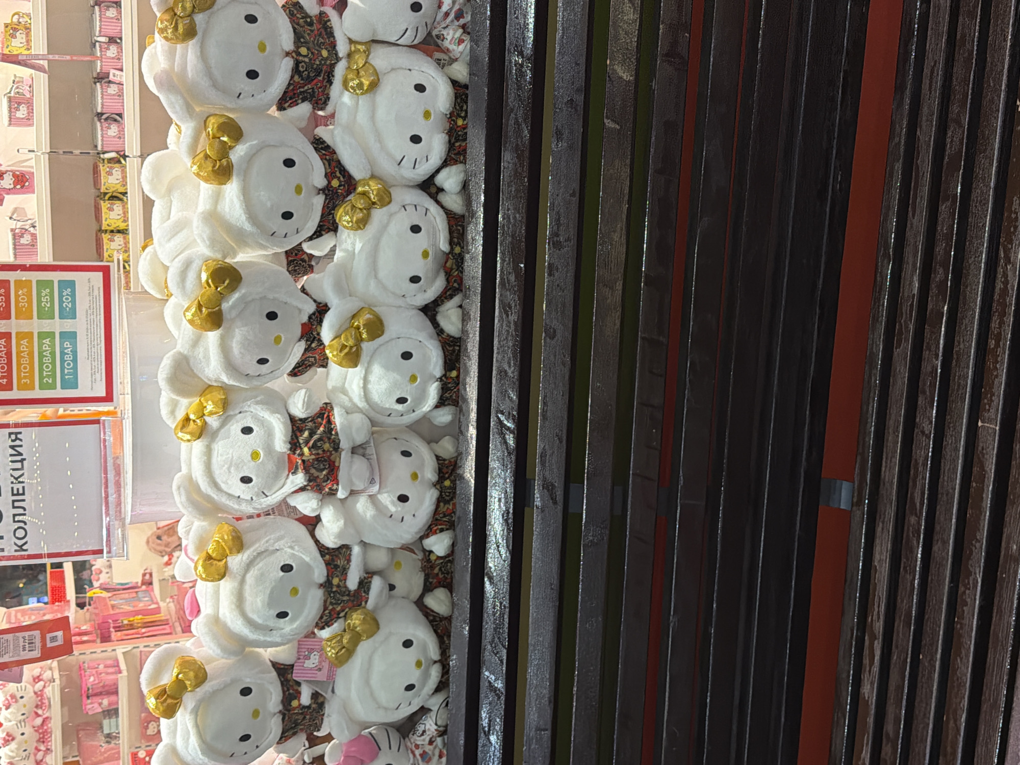 The image shows a row of stuffed Hello Kitty toys displayed on a dark, wooden shelf. The toys are white with hoods and have gold bows. They are wearing patterned vests. In the background, shelves are stocked with various Hello Kitty merchandise, including mugs and other plush toys. A sign with Russian text is also visible. The lighting suggests an indoor retail environment.