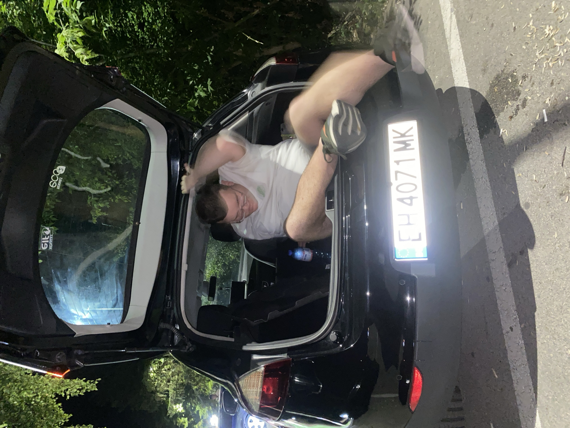 The image shows a person with glasses, wearing a white t-shirt and grey shorts, sitting in the open trunk of a black car. The person is in motion, with their right arm raised and their legs extended. A blue bottle of water is visible on the floor of the trunk. The car's license plate, "EH 4071 MK", is illuminated. The scene appears to be outdoors at night, with trees and foliage visible in the background. The ground is paved, with tire marks and some fallen leaves. The overall impression is of a candid, informal moment. The license plate suggests the location is in Bulgaria. Without further information, it is impossible to definitively determine the specific location within Pleven, Bulgaria.
