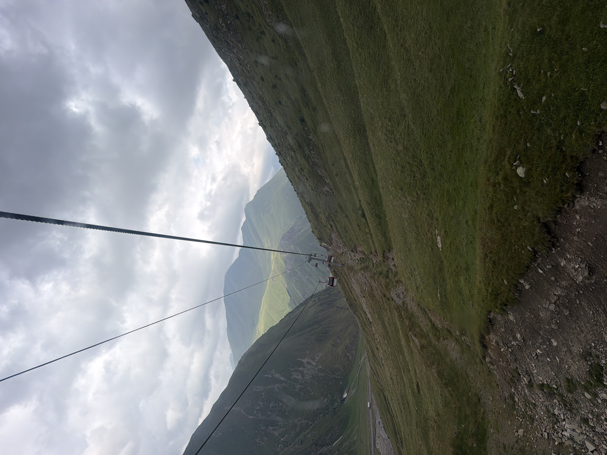 A cable car ascends a grassy mountain slope under a cloudy sky. Two red and white cable car cabins are visible on the right side of the frame, suspended from a thick cable. The mountain is covered in green grass and rocks, with some patches of bare earth. In the distance, more mountains are visible, some appearing a lighter shade of green, indicating they are further away or perhaps covered in a different type of vegetation. A road can be seen in the valley below on the left, with a small white vehicle on it. The overall impression is a natural, mountainous landscape, likely a popular tourist destination given the cable car. The location is in the vicinity of Almasiani, Georgia.