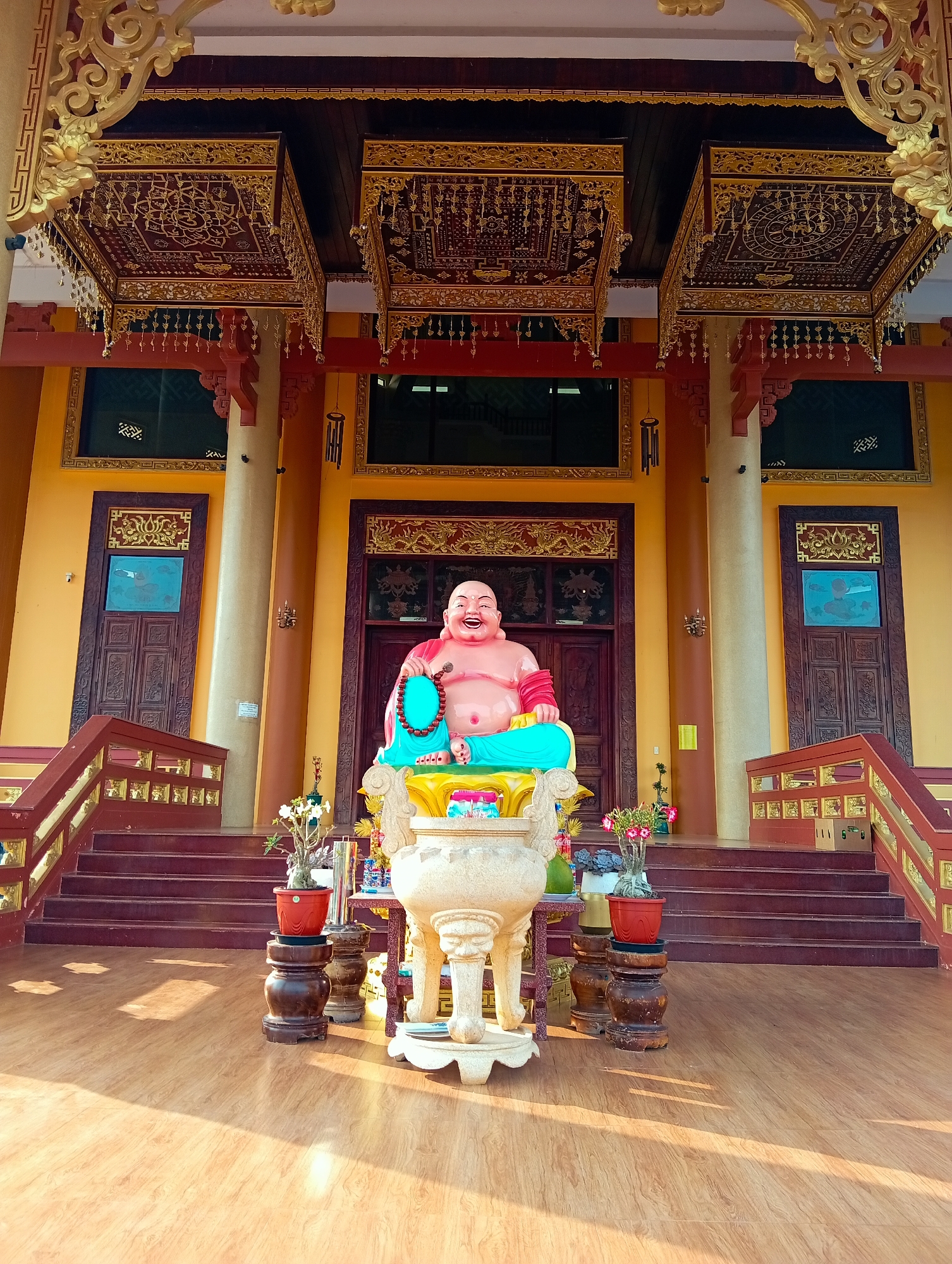 This vibrant image depicts the ornate entrance of a temple or pagoda in Phú An, Vietnam, under bright sunlight, suggesting either morning or late afternoon.
At the center, a large, smiling statue of Budai, often known as the Laughing Buddha, is prominently displayed. The statue features pink skin, a red top, and blue lower garments, holding a teal beaded mala in its left hand. It is seated on a golden pedestal, which rests upon a large, intricately carved stone censer. Surrounding the base of the statue and the censer are various potted plants, some with flowering desert roses, and other small decorative items or offerings.
The building behind the statue is a striking yellow, richly adorned with red and gold architectural details. Two elaborate, canopy-like structures, featuring intricate gold patterns on a dark red background and dangling golden ornaments, hang from the ceiling above the main entrance. Flanking the central display are two dark wooden doors with carved panels and decorative gold architraves, topped by dark windows. Two substantial, light-colored columns rise from the ground, supporting the structure.
Wide, dark red steps with ornate wooden railings, highlighted with gold accents, lead up to the temple entrance. The foreground reveals a light wooden or tiled floor, upon which the sun casts distinct shadows. The scene is devoid of people, conveying a peaceful and sacred atmosphere. No discernible text is visible in the image.