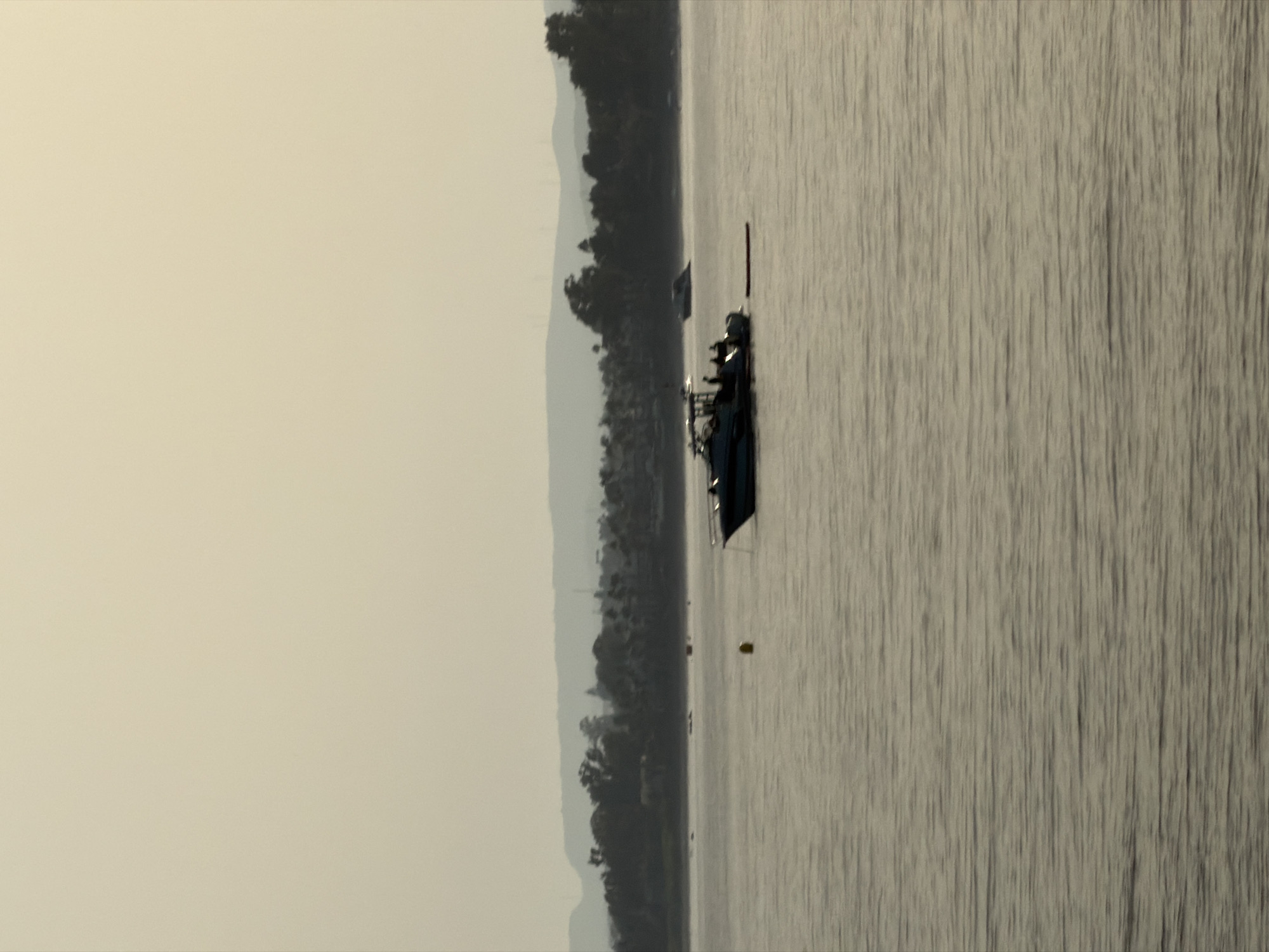This is a vertical, eye-level photograph of a body of water, possibly the Mediterranean Sea, near Estepona, Spain, at dusk. The sky is a hazy, pale yellow, with distant, silhouetted hills. The water is calm with subtle ripples. In the foreground, on the right, a dark-hulled boat with a light-colored superstructure is partially visible. Further to the left, a larger, white boat with a dark trim is in the center of the frame. This boat has several people visible on its deck, and a red object floats in the water behind it. A yellow buoy is visible in the water between the two boats. In the background, a line of trees and buildings is silhouetted against the horizon, suggesting a coastal town or city. The overall impression is one of tranquility and calm before nightfall.