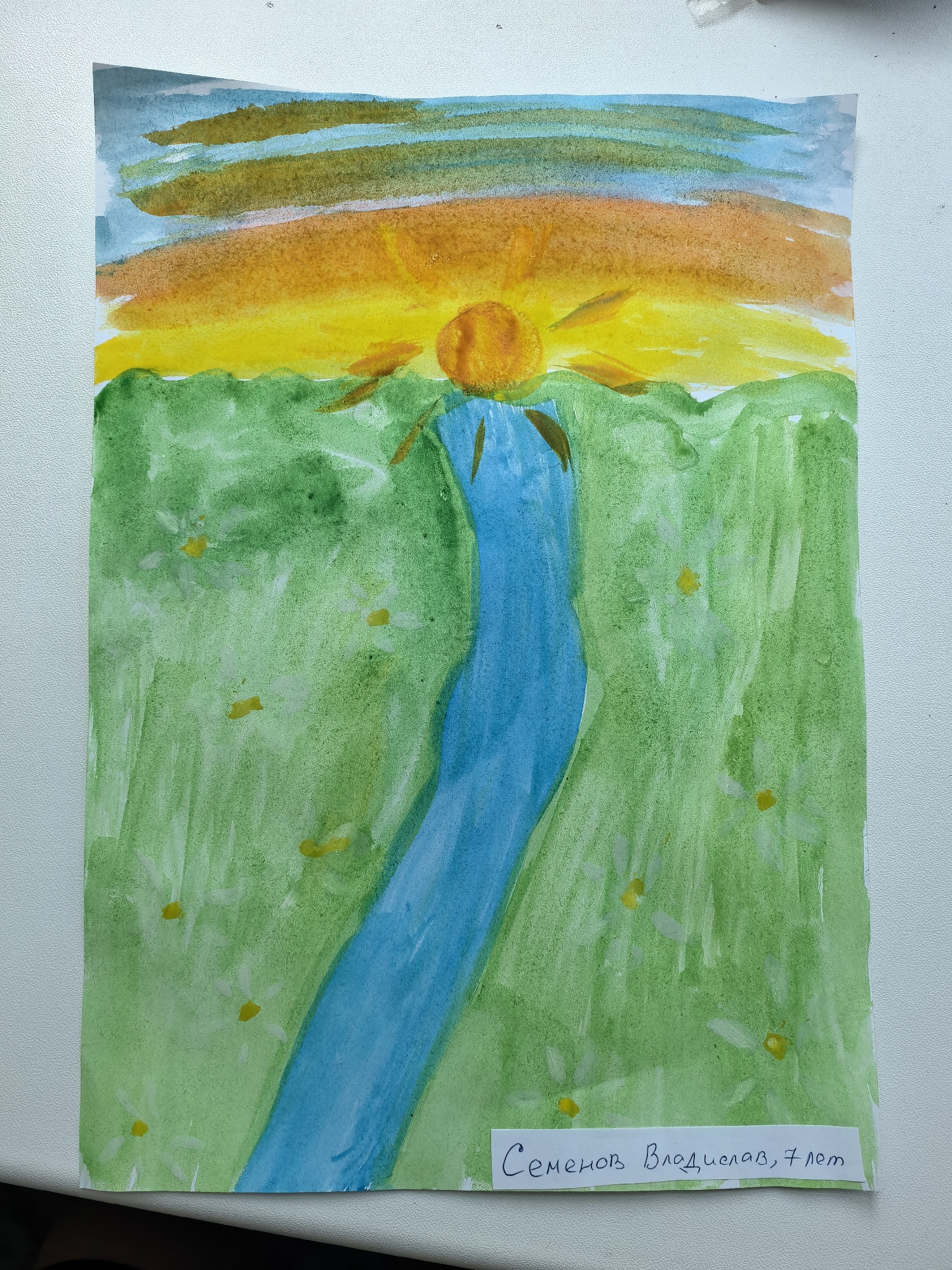 A child's watercolor painting depicts a landscape scene. A blue river flows vertically through a green field dotted with small white flowers. Above the field, a yellow and orange sunset with a visible sun ball is painted across the sky with light blue streaks. A white label at the bottom of the painting contains text in Cyrillic which translates to "Semenov Vladislav, 7 years old," suggesting the age and name of the artist. The location, Sterlitamak, Russia is not visibly indicated in the painting.