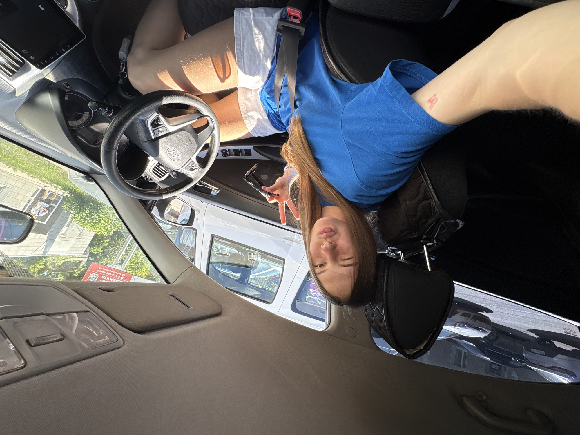A young woman with blonde hair, wearing a blue t-shirt and white shorts, sits in the driver's seat of a car and poses for a selfie. She is winking and holding up two fingers in a peace sign. A small butterfly tattoo is visible on her left arm. Outside the car, parked vehicles and buildings can be seen. The scene appears to be in a residential area, likely in the city of Perm, Russia, given the presence of Russian text on a sign visible through the windshield. The car's interior features a steering wheel, dashboard, and infotainment screen. Sunlight streams into the car, creating strong shadows.