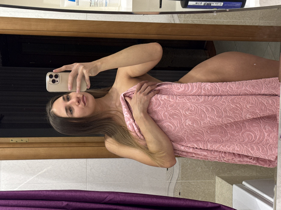A woman in a bathroom setting is taking a selfie in a mirror. She is partially covered with a large pink towel. The phone in her hand is a beige or gold color. The background includes bathroom fixtures, a wooden cabinet, and various toiletries on a shelf. The lighting is soft, and the woman's expression is neutral.