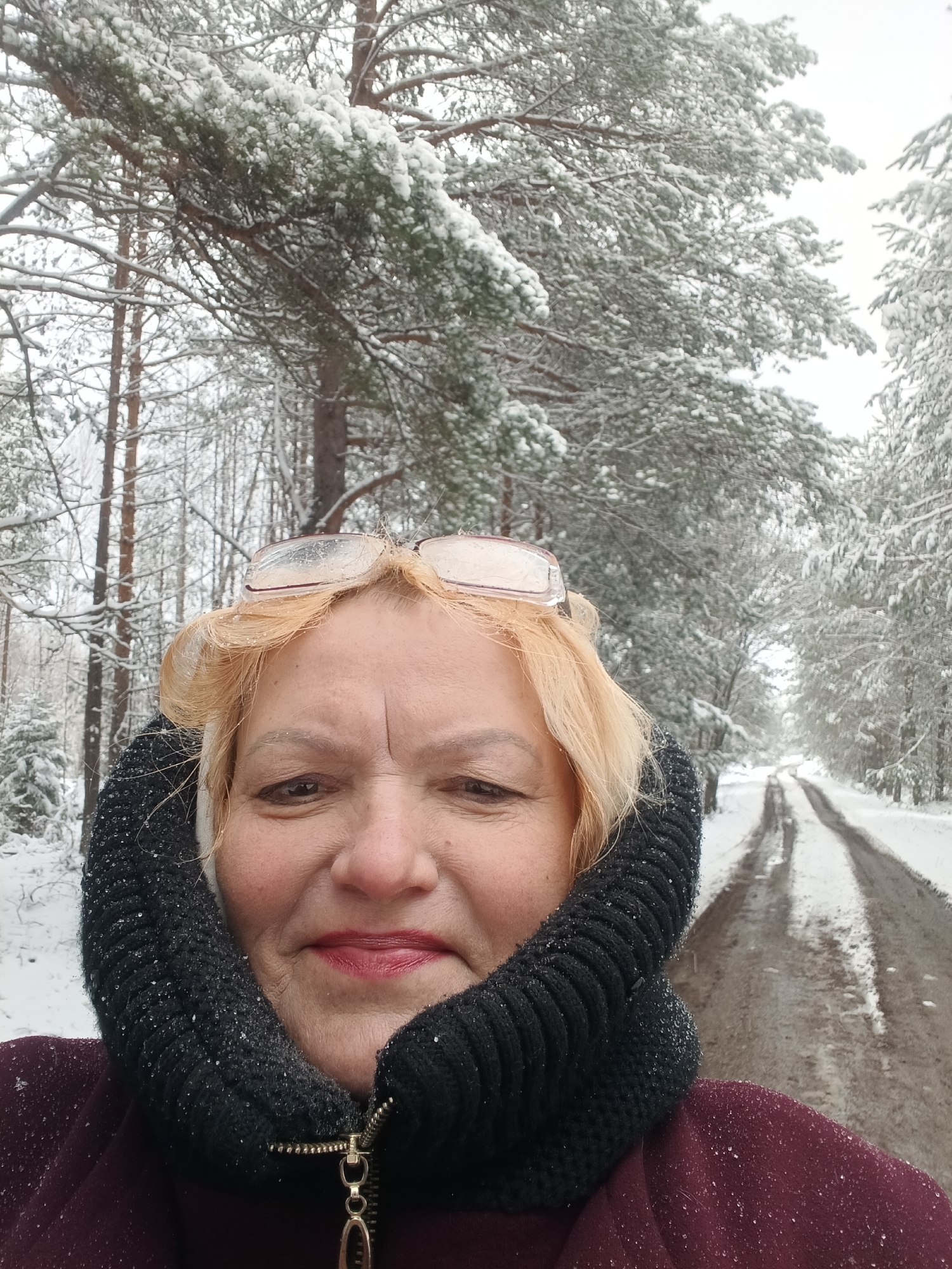The image shows a woman in an outdoor winter scene. She is wearing a dark-colored, knitted scarf around her neck and a maroon jacket. Her blond hair is visible above the scarf and she has glasses perched on top of her head. The woman is smiling slightly.
The background features a snowy forest scene. A dirt road with two distinct tire tracks runs through the center of the forest, leading into the distance. The trees are covered in snow, creating a wintry atmosphere. Given the context of Красный Маяк, Russia, it can be inferred that this is a forest or park area in or near the city, covered in snow during winter.