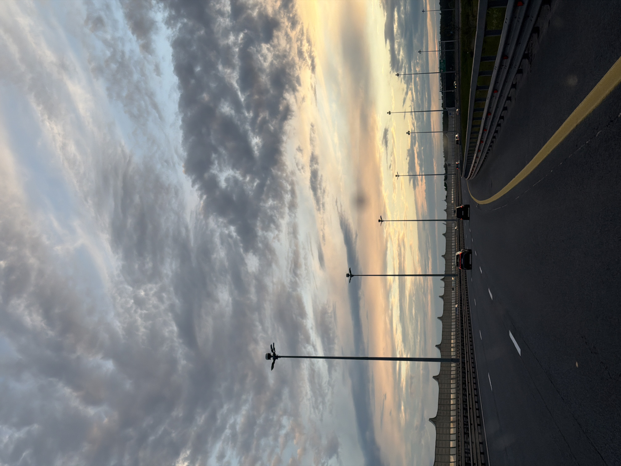 The image captures a wide, multi-lane highway at either sunrise or sunset, set against a dramatic, cloudy sky. The perspective is from the road itself, looking forward as it stretches into the distance.
The highway is paved with dark asphalt, featuring clear white dashed lane markers and a prominent solid yellow line that likely delineates a lane or shoulder. On the right side, metal guardrails are visible, while a tall sound barrier or protective wall runs along the left. A series of modern, tall streetlights, each with multiple fixtures, are spaced evenly on both sides of the road, creating a strong sense of depth.
Several vehicles, appearing as dark silhouettes against the brighter horizon, are seen in the distance, traveling away from the viewer. The sky dominates the upper portion of the image, filled with a dynamic mix of dark, textured grey clouds and lighter, more wispy formations. Patches of warm, golden light pierce through the clouds near the horizon, indicating the "golden hour" and casting a soft, transitioning glow over the scene. In the far distance, beyond the left-hand barrier, faint outlines of dark vegetation or a forest are visible.
This scene depicts an expressway in Saint Petersburg, Russia, during a time of day characterized by striking natural light and ongoing vehicular movement. There is no visible text in the image.