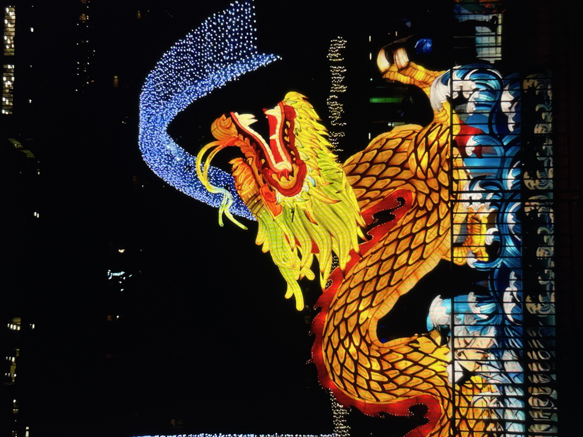 A nighttime shot shows a large illuminated dragon display on the side of a building in an unknown city. The dragon's head is yellow and green with a red mouth and yellow whiskers. The body of the dragon is gold with red fins. A blue arc of lights appears to emanate from behind the dragon's head. The background is dark, with some other building lights visible. The building on which the dragon is displayed has blue and white decorations alongside the dragon.